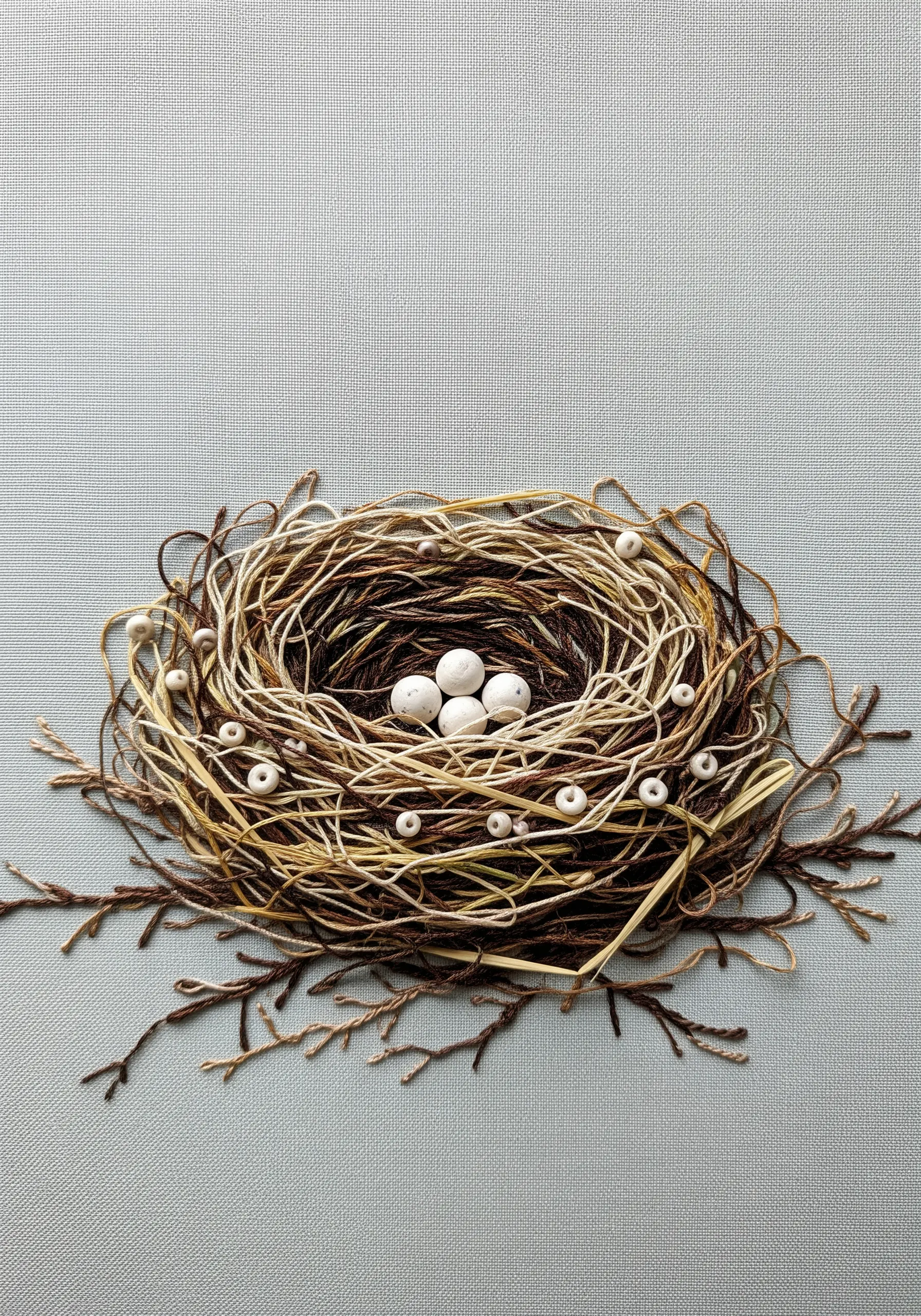A highly textural embroidered bird's nest made from layered brown and cream threads, with white beads as eggs.