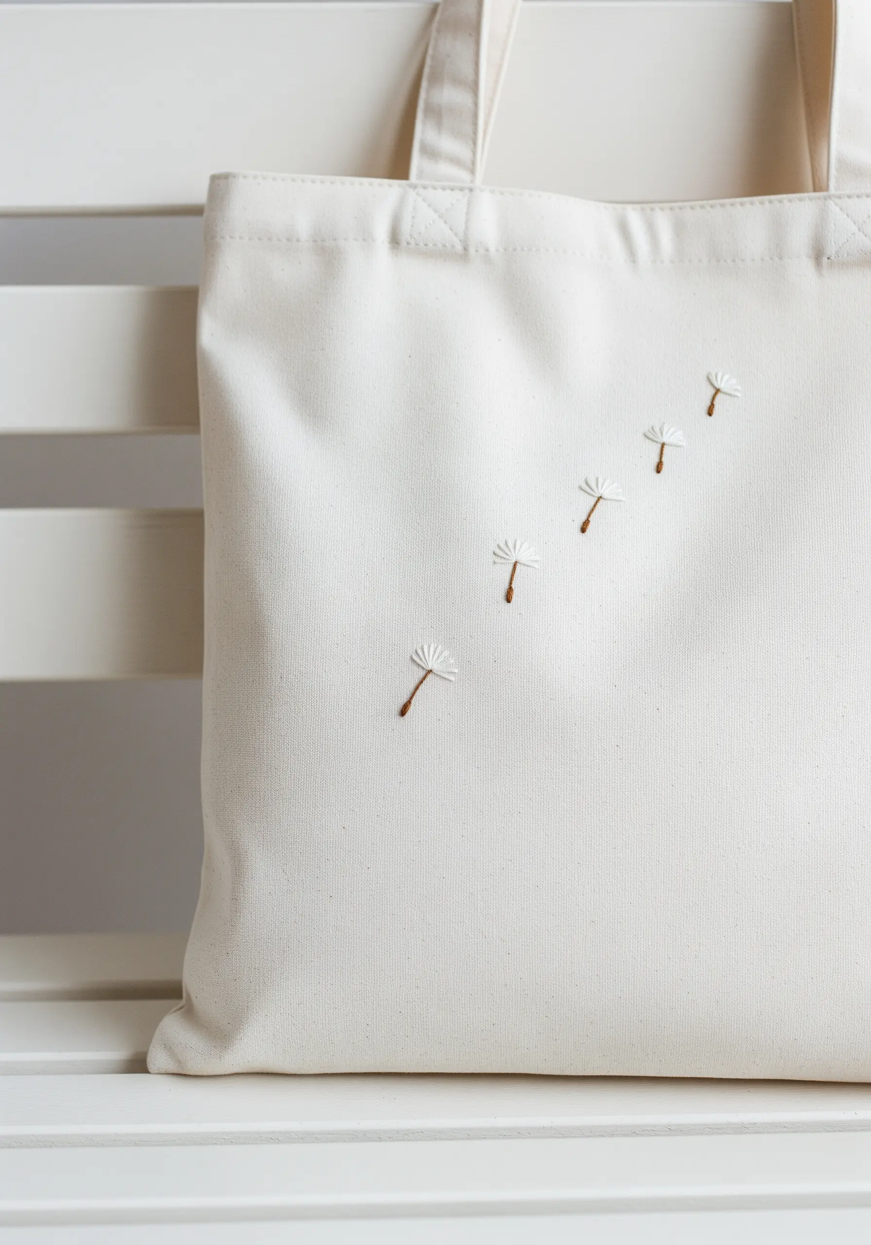A trail of five delicate embroidered dandelion seeds floating across a white tote bag.