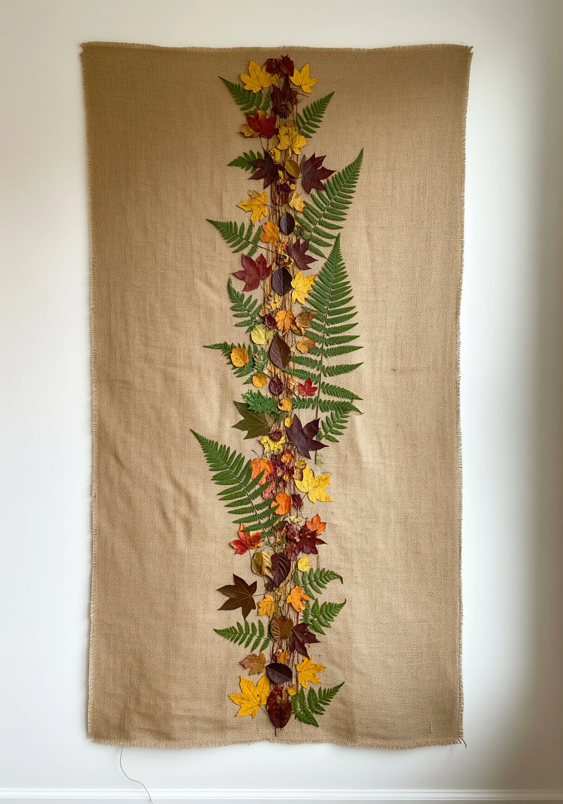 A vertical garland of pressed autumn leaves and ferns stitched onto a large burlap wall hanging.