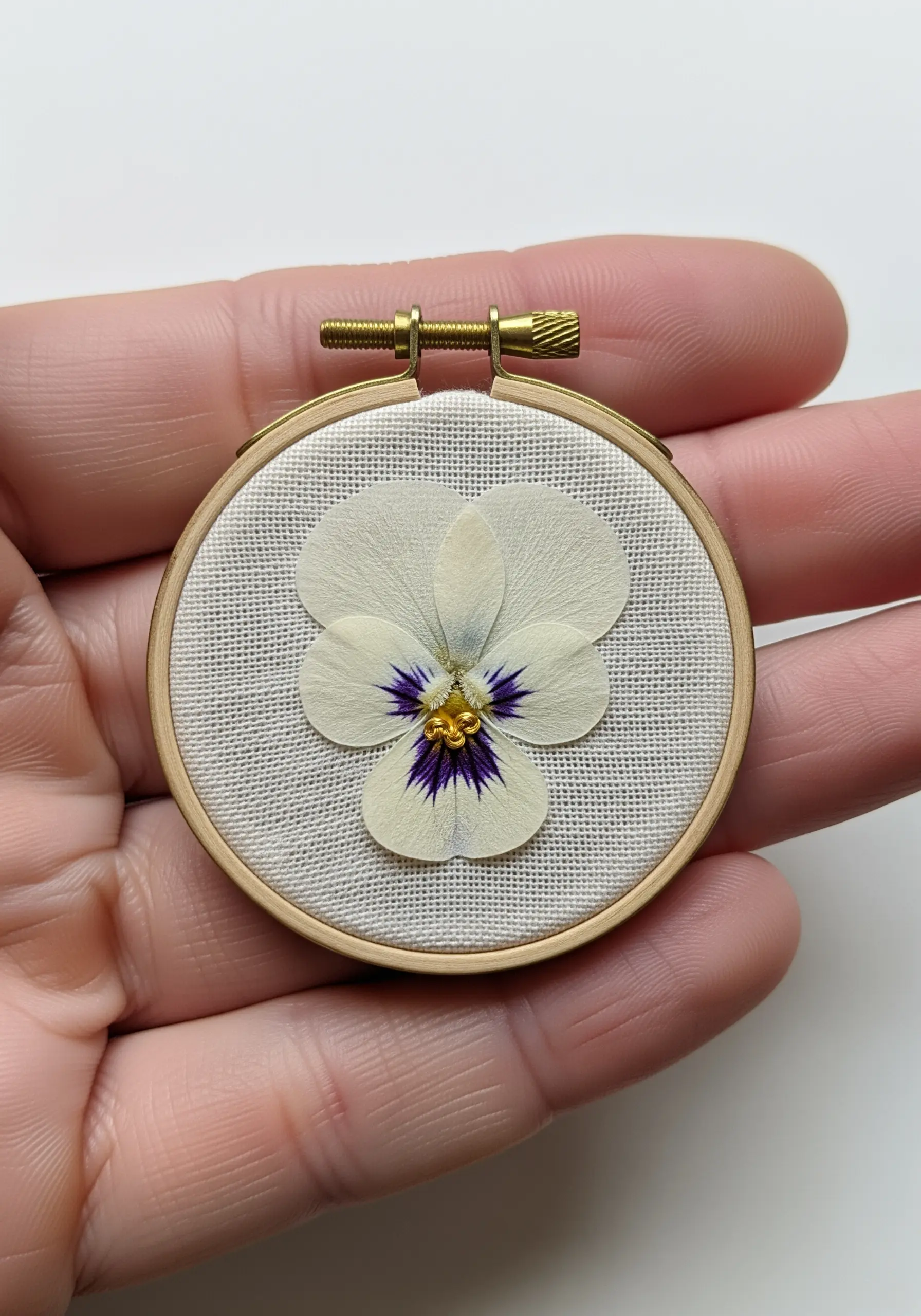 A tiny embroidery hoop held in hand, featuring a single white pansy as a wearable pendant.