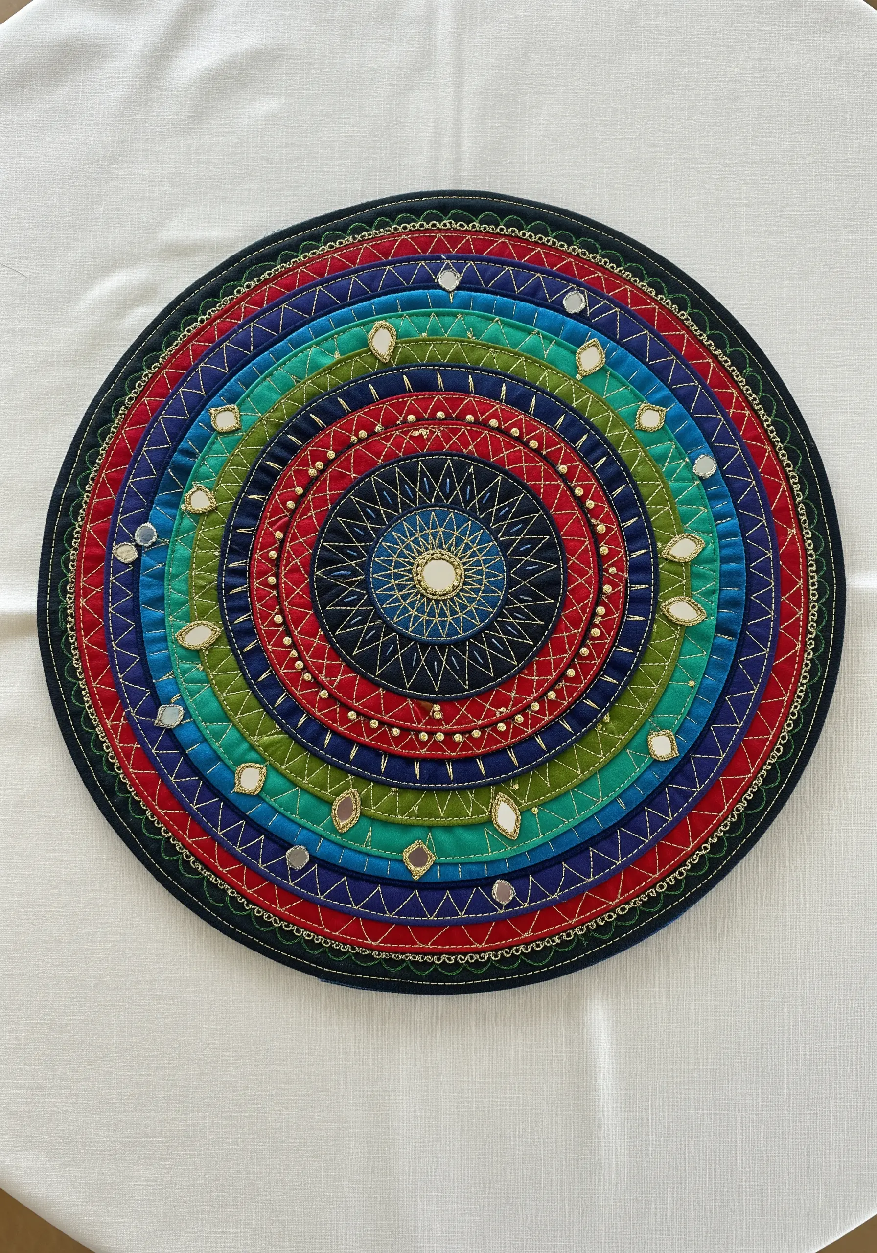 A circular, multicolored fabric mandala placemat with intricate machine stitching, beads, and mirrors.