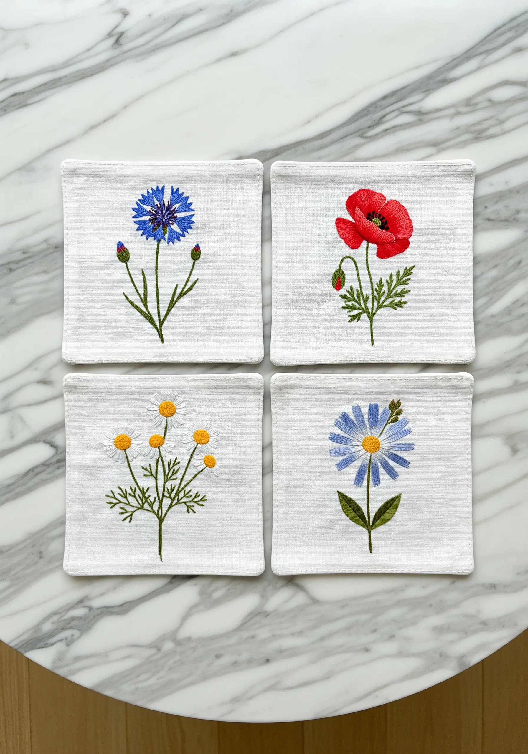 Four white embroidered coasters on a marble table, each with a different wildflower.
