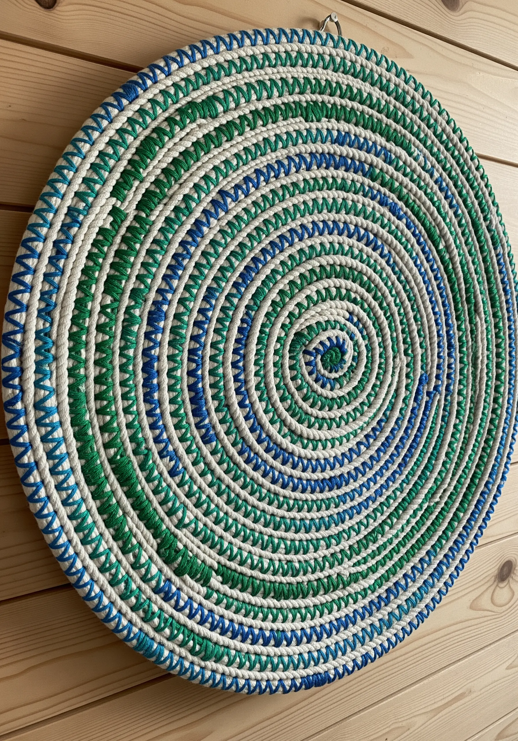 A circular wall hanging made of coiled white rope wrapped in blue and green thread.