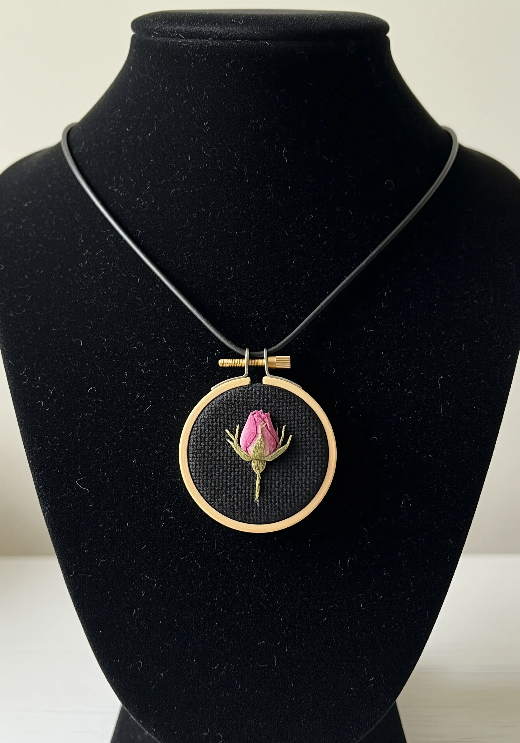 A miniature embroidery hoop necklace featuring a tiny pressed rosebud on black fabric.