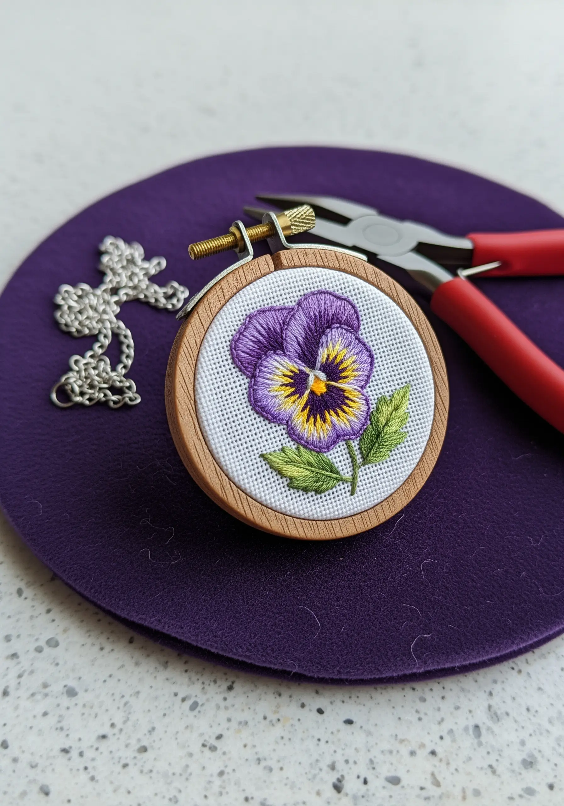 A tiny embroidered pansy set in a miniature hoop pendant, next to jewelry-making tools.