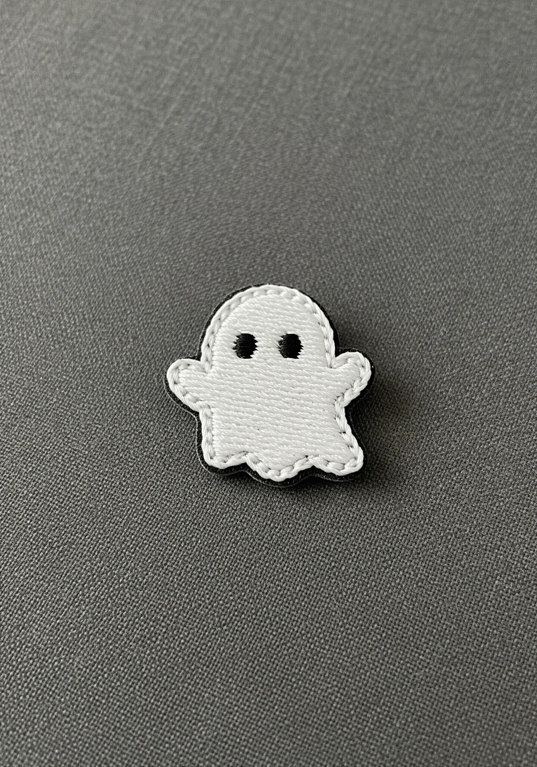 A small, friendly ghost embroidered as a patch with a black outline.
