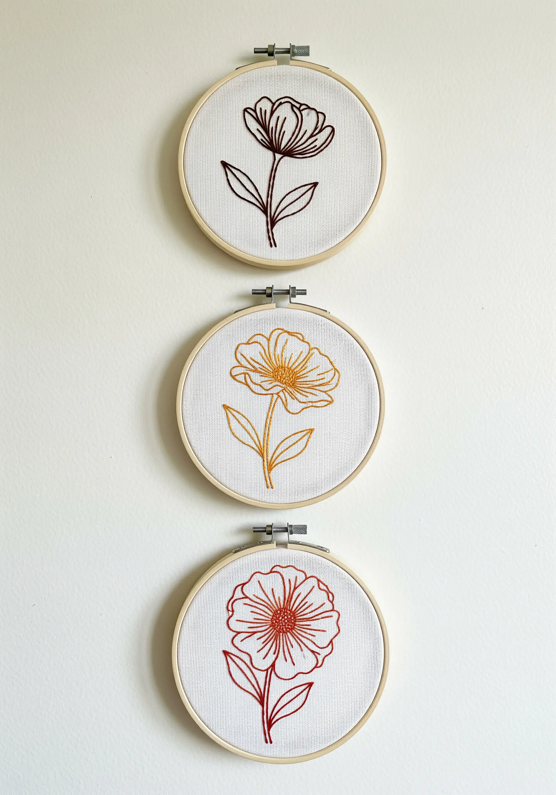 A trio of small embroidery hoops, each with a single line art flower in a warm color.