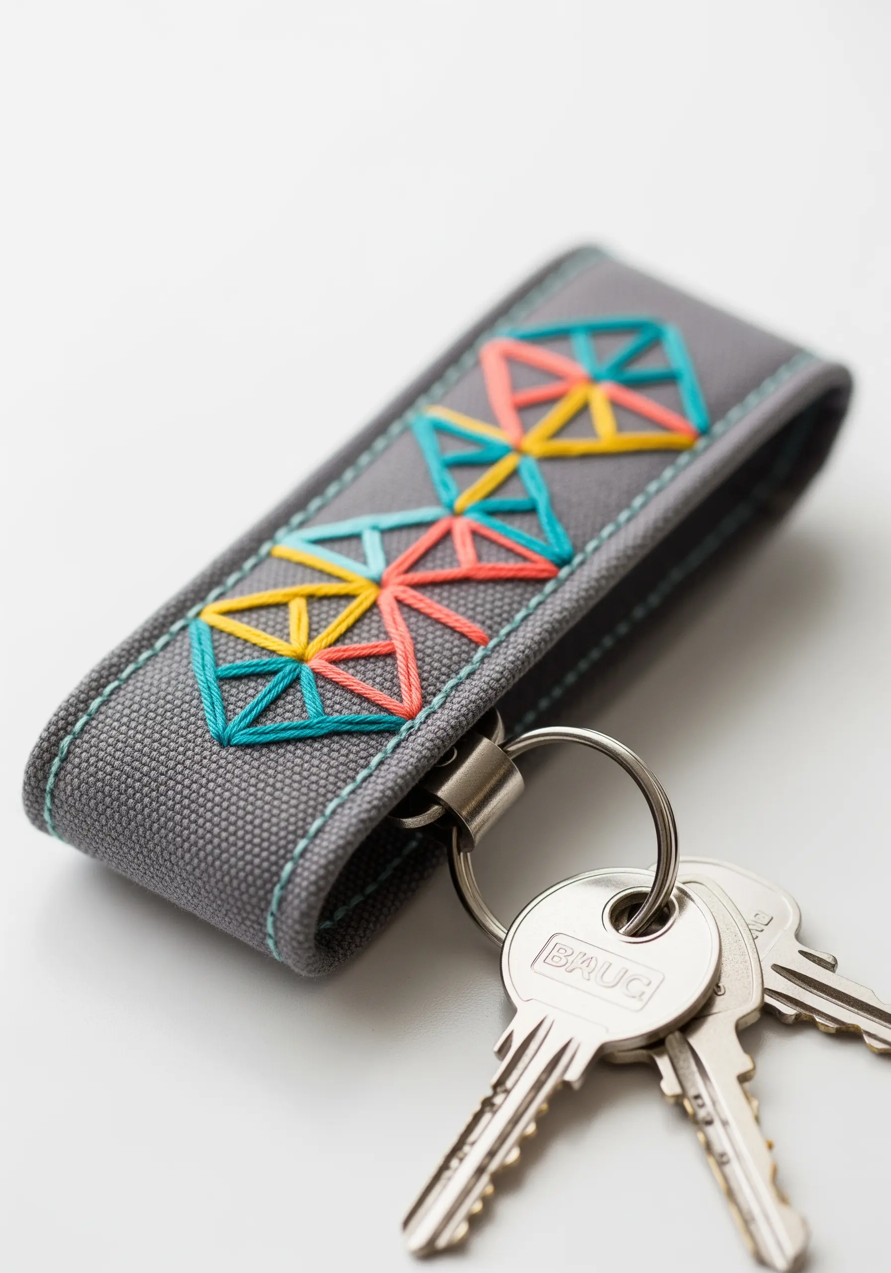 A gray fabric keychain with a colorful geometric pattern stitched in teal, orange, and yellow.