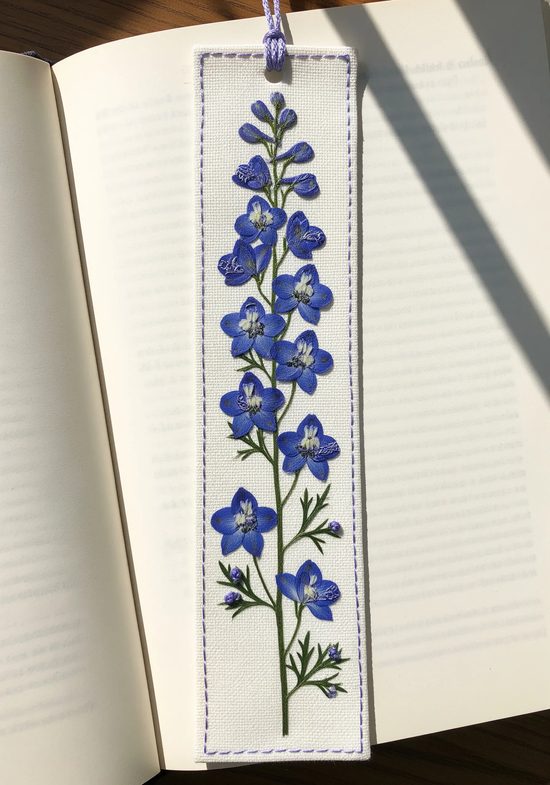 A bookmark featuring a delphinium stalk made from pressed flowers and photorealistic embroidery.