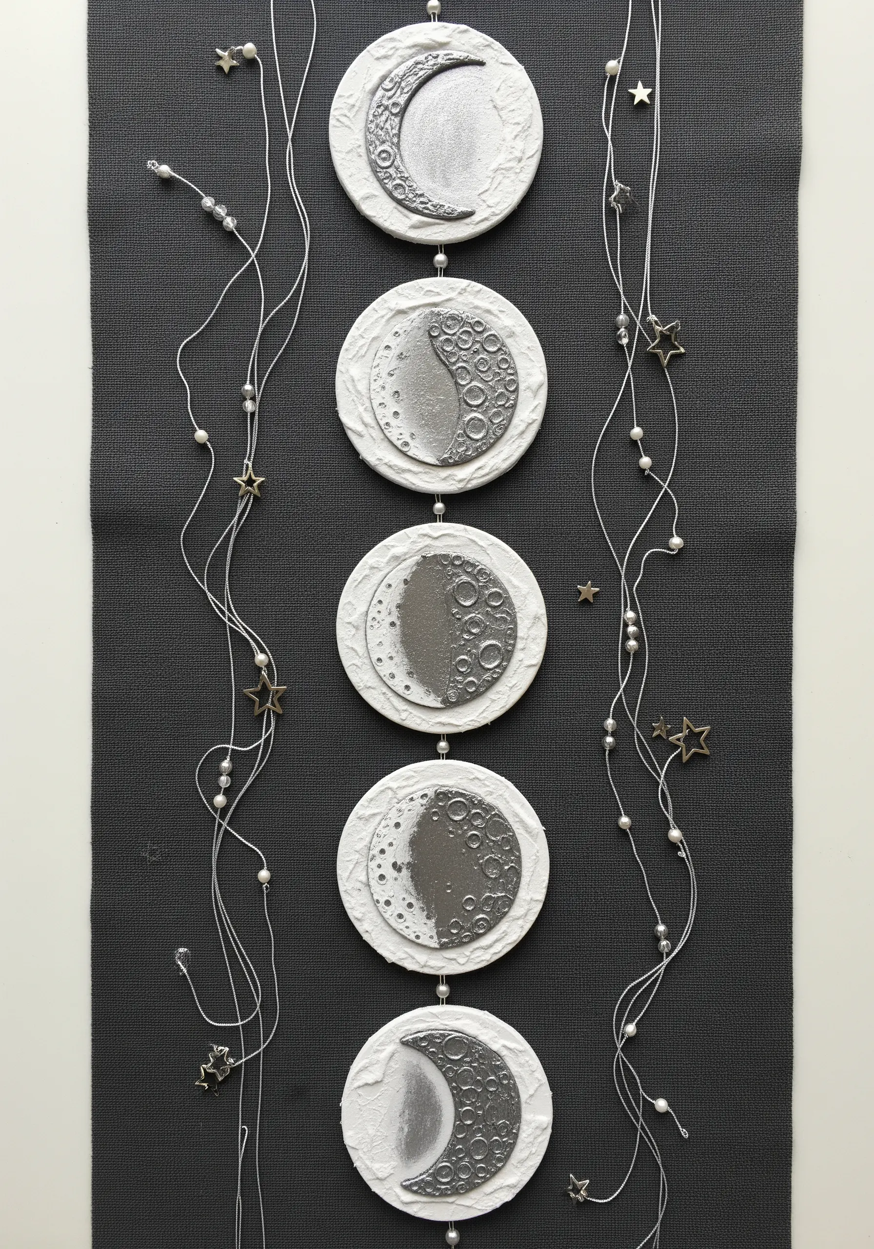 A vertical wall hanging showing the phases of the moon made from textured clay discs and beads.