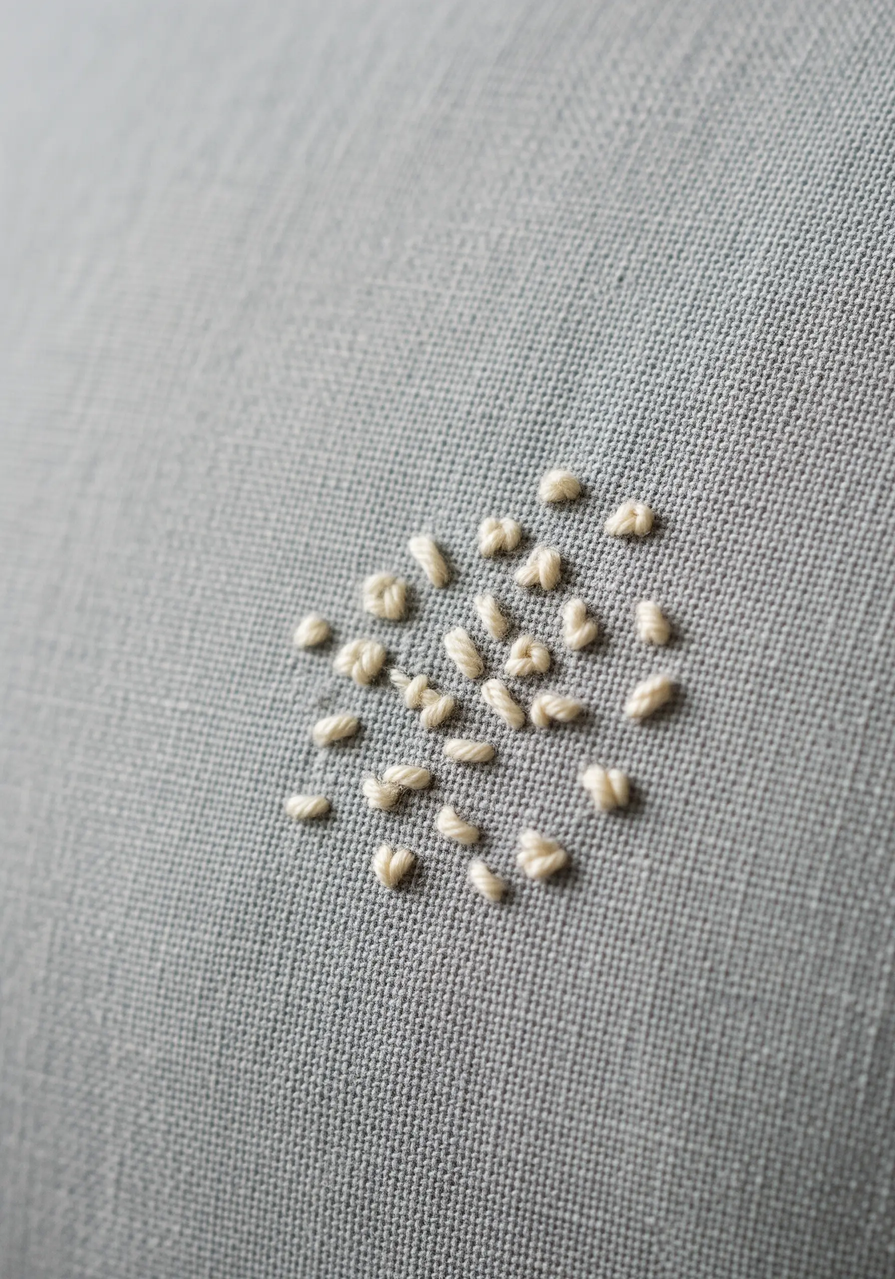 A small, circular cluster of ivory French knots on a light gray-blue linen pillowcase.
