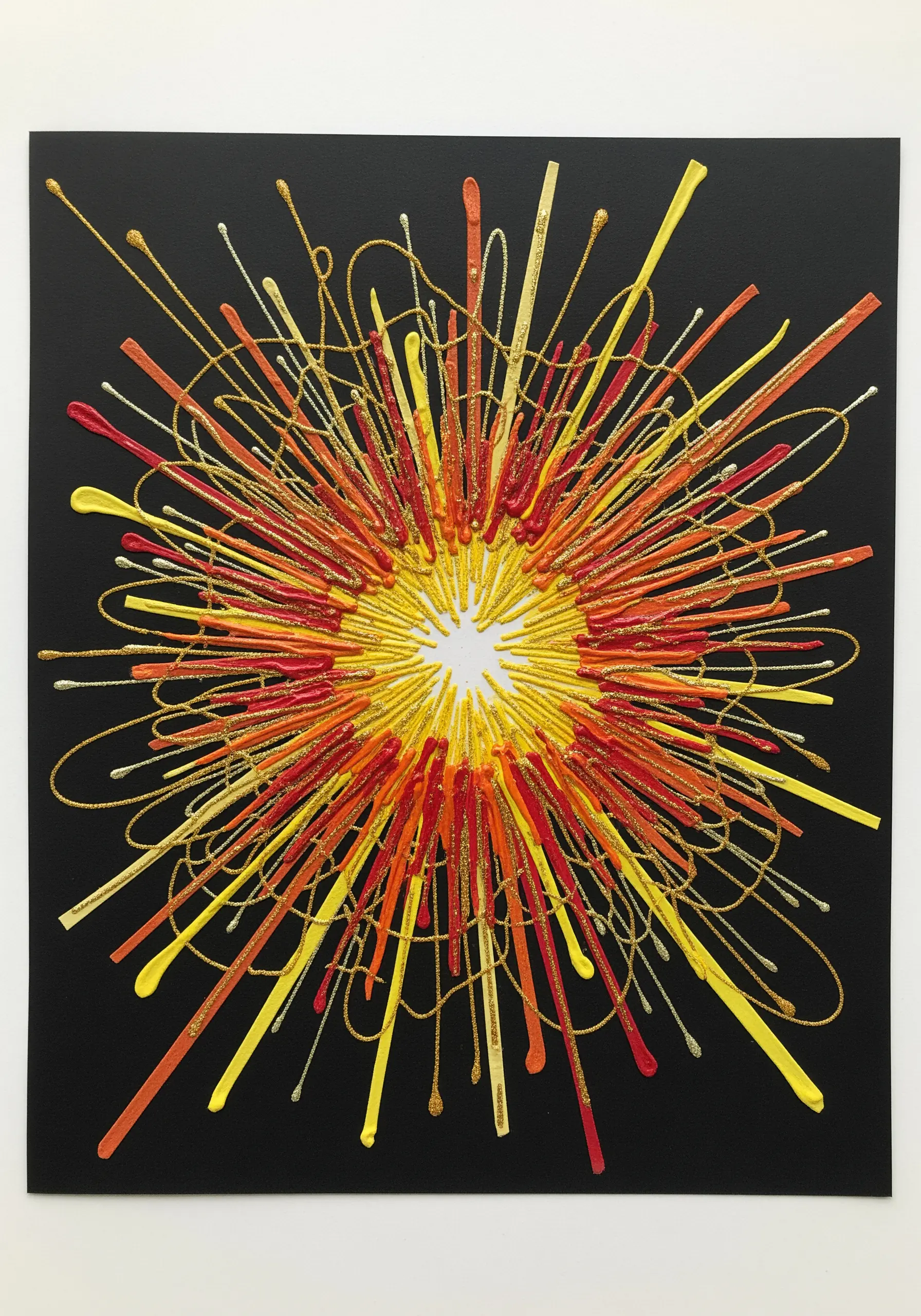 An abstract burst of color made with yellow, orange, and red paint, glitter, and thread on black paper.