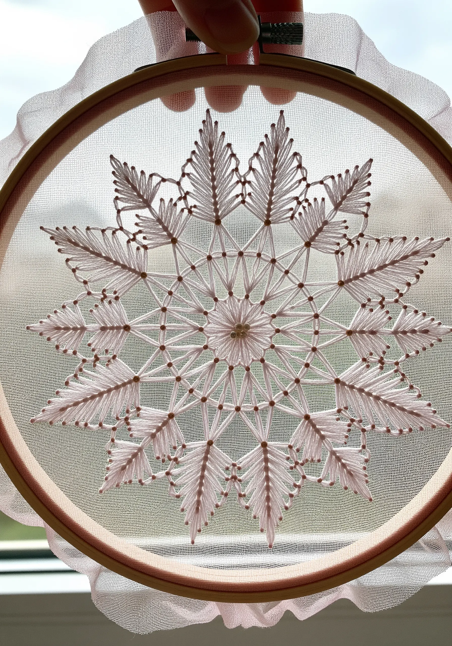 Delicate white snowflake mandala embroidered on sheer pink organza, held up to a window.