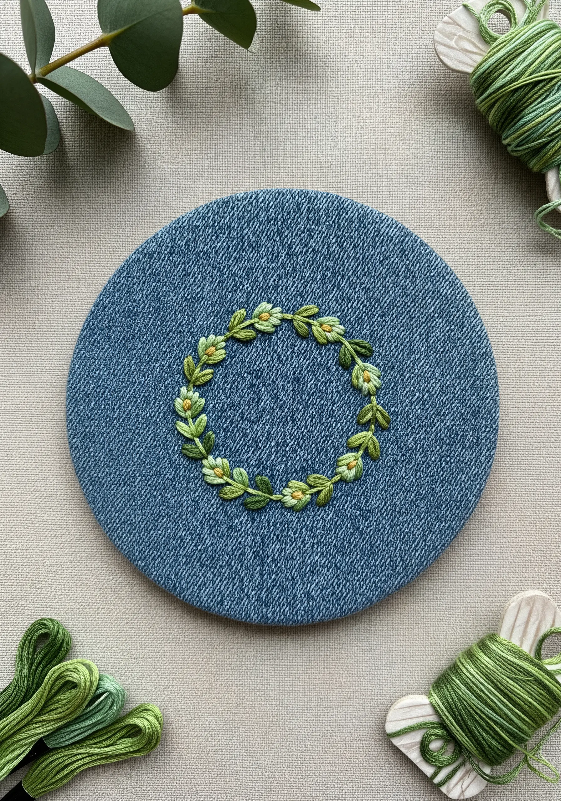 A delicate wreath of green leaves and small white flowers stitched on blue denim in a hoop.