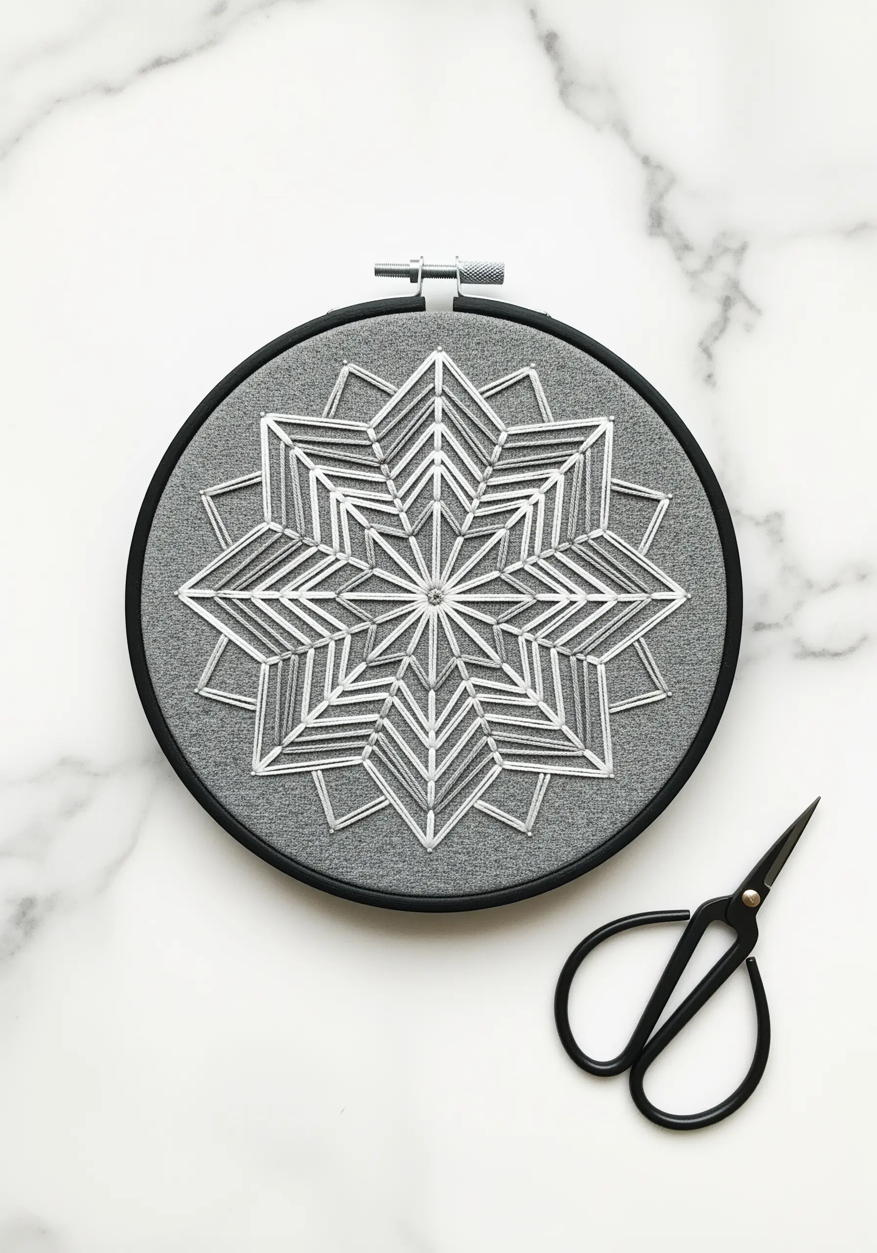 Geometric snowflake mandala stitched in white thread on gray fabric in a black hoop.