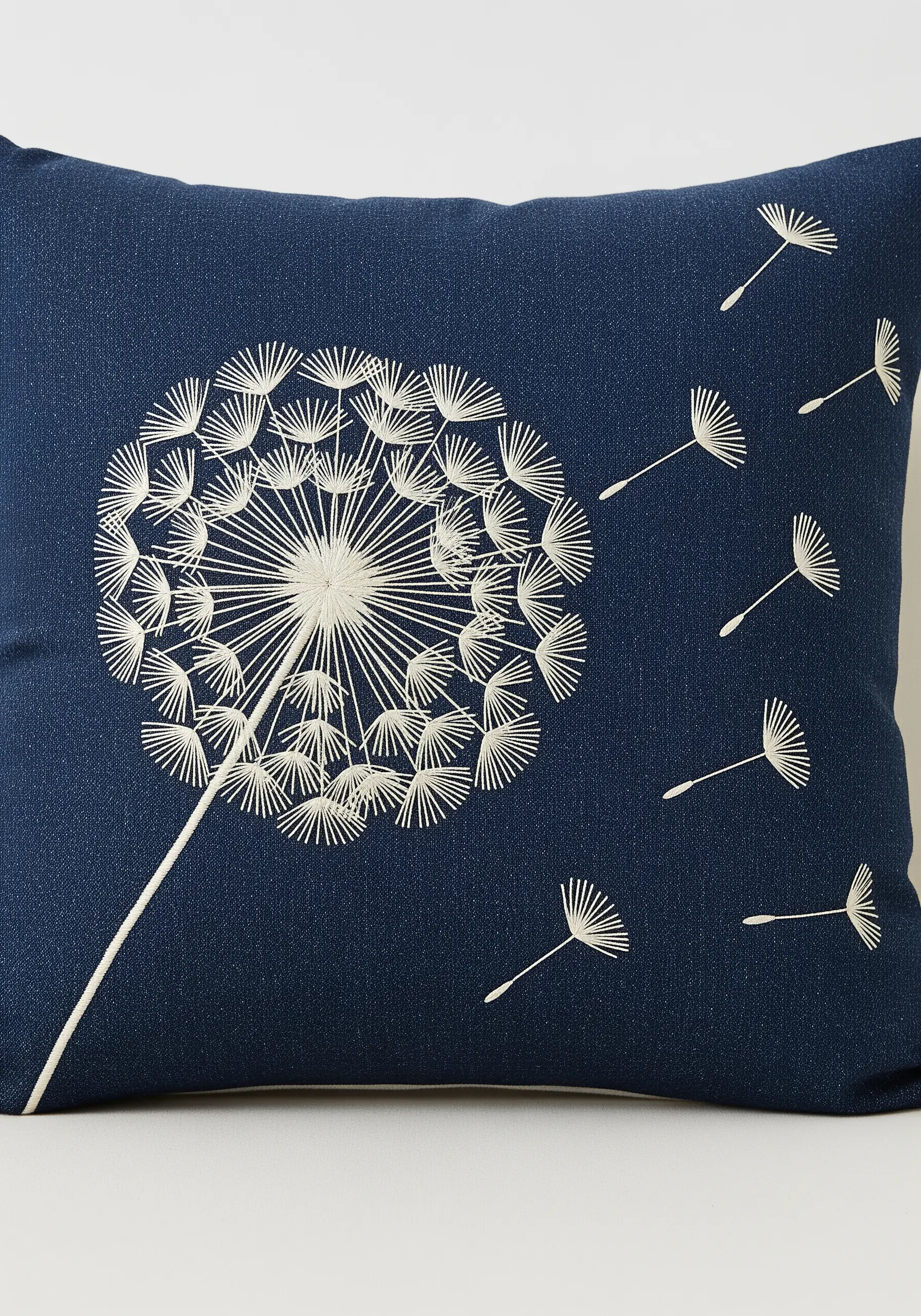 A white embroidered dandelion with seeds blowing in the wind on a dark blue cushion cover.