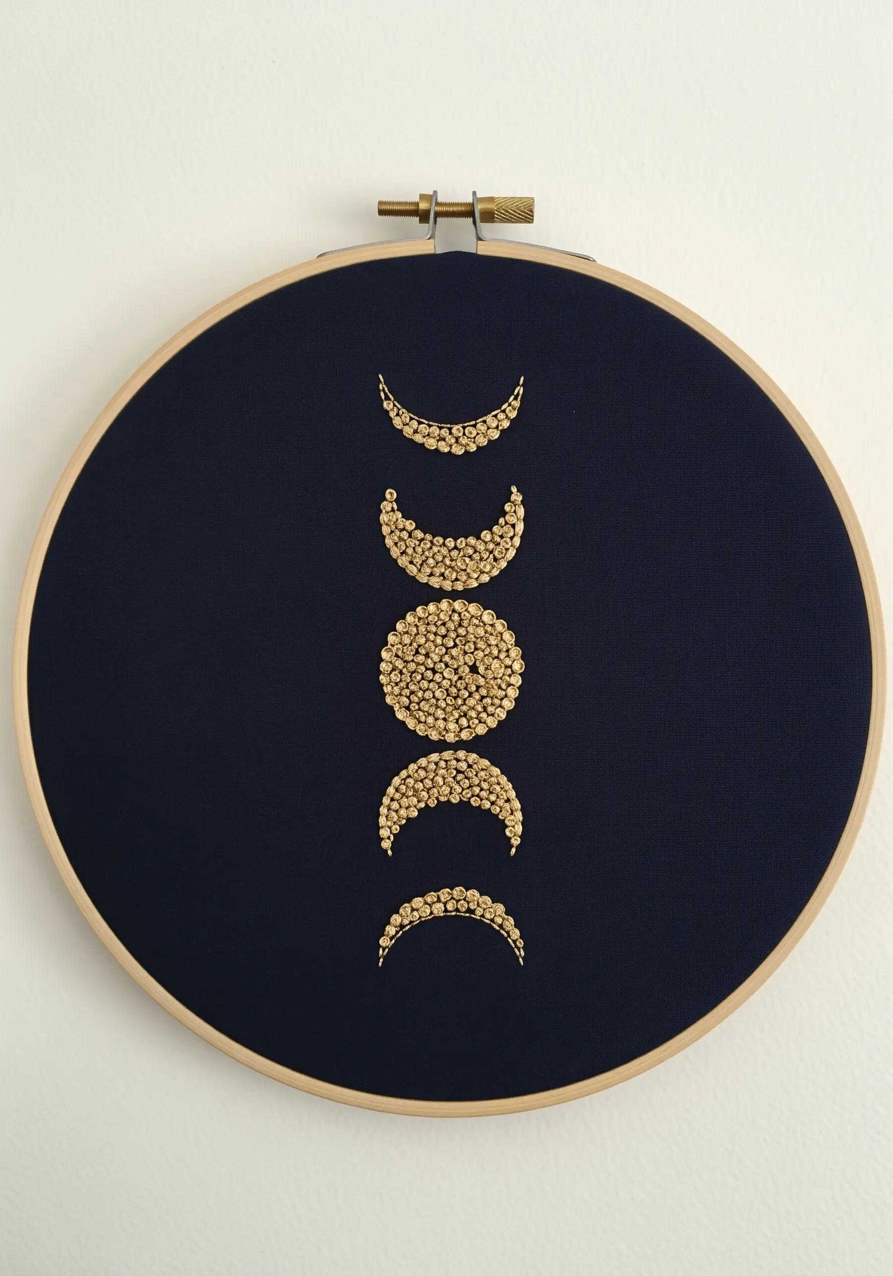 Moon phases design in a hoop, filled entirely with shimmering gold seed beads on navy fabric.