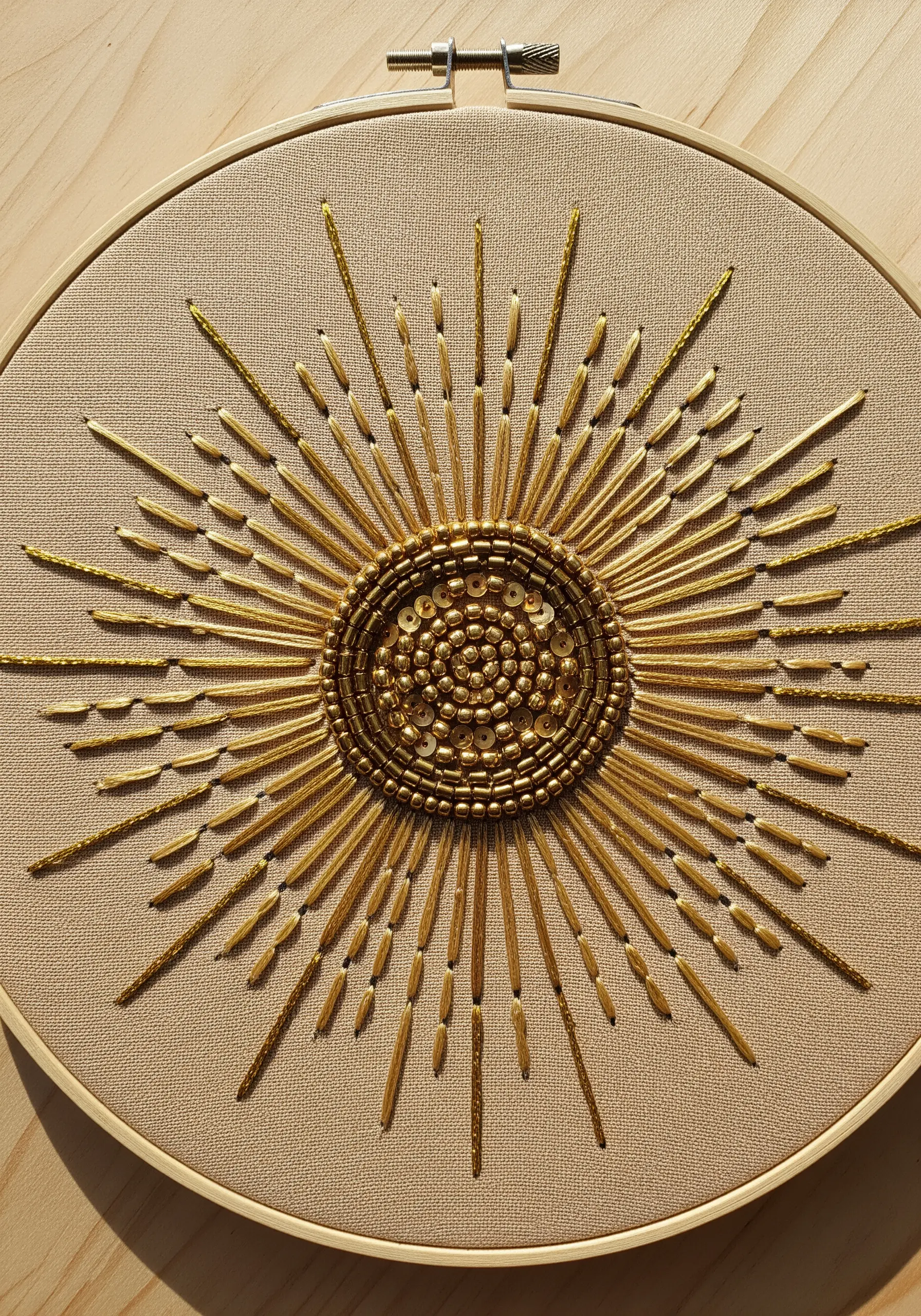 A sunburst motif embroidered with gold bugle beads, seed beads, and sequins on neutral fabric.