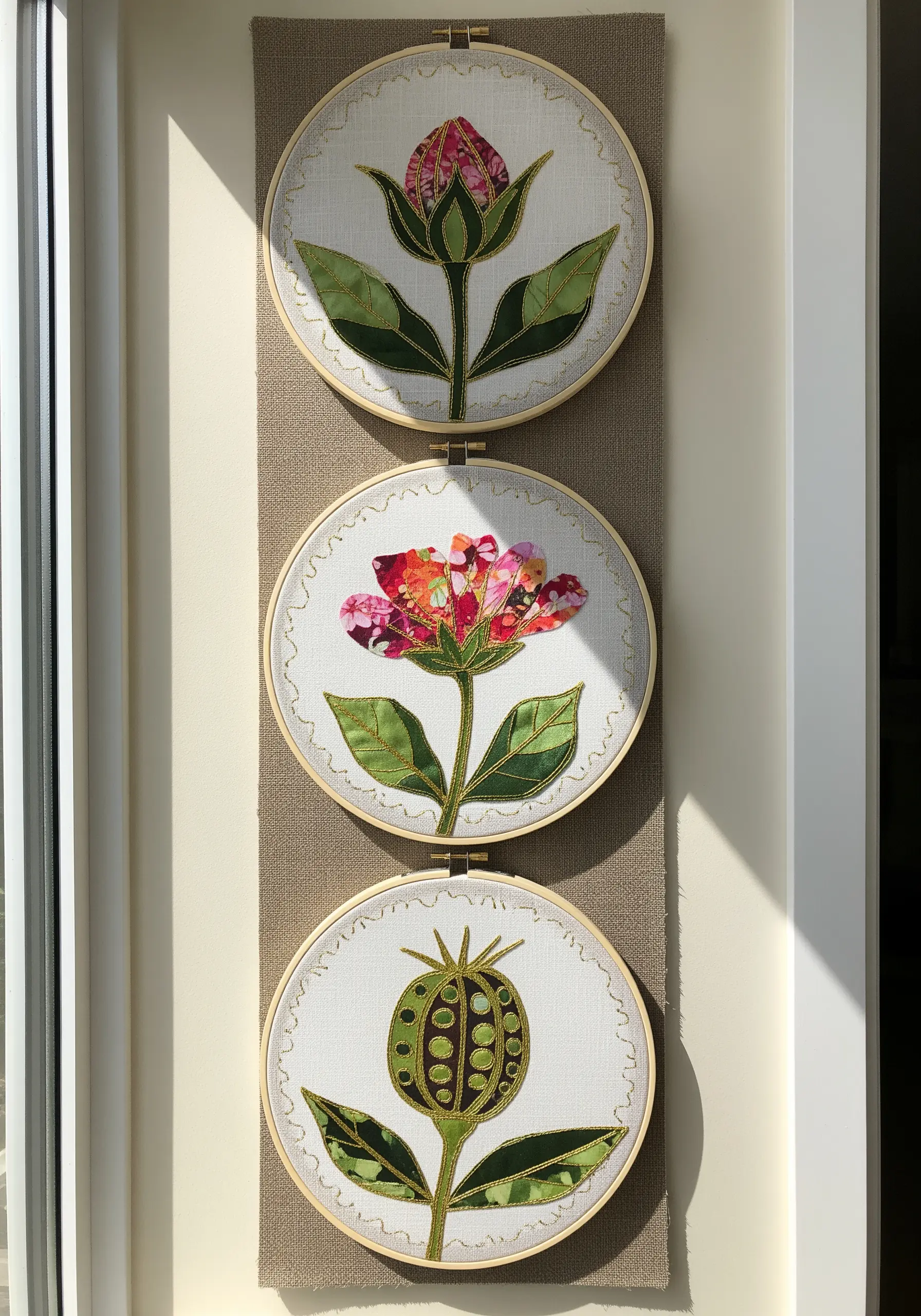 Three embroidery hoops displaying floral appliqué with gold thread outlines, resembling stained glass.