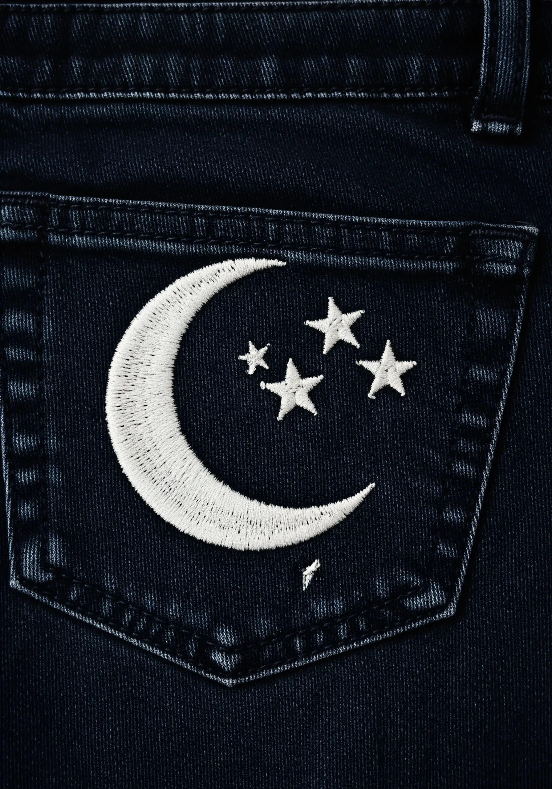 A crescent moon and stars embroidered in bright white satin stitch on a dark denim pocket.