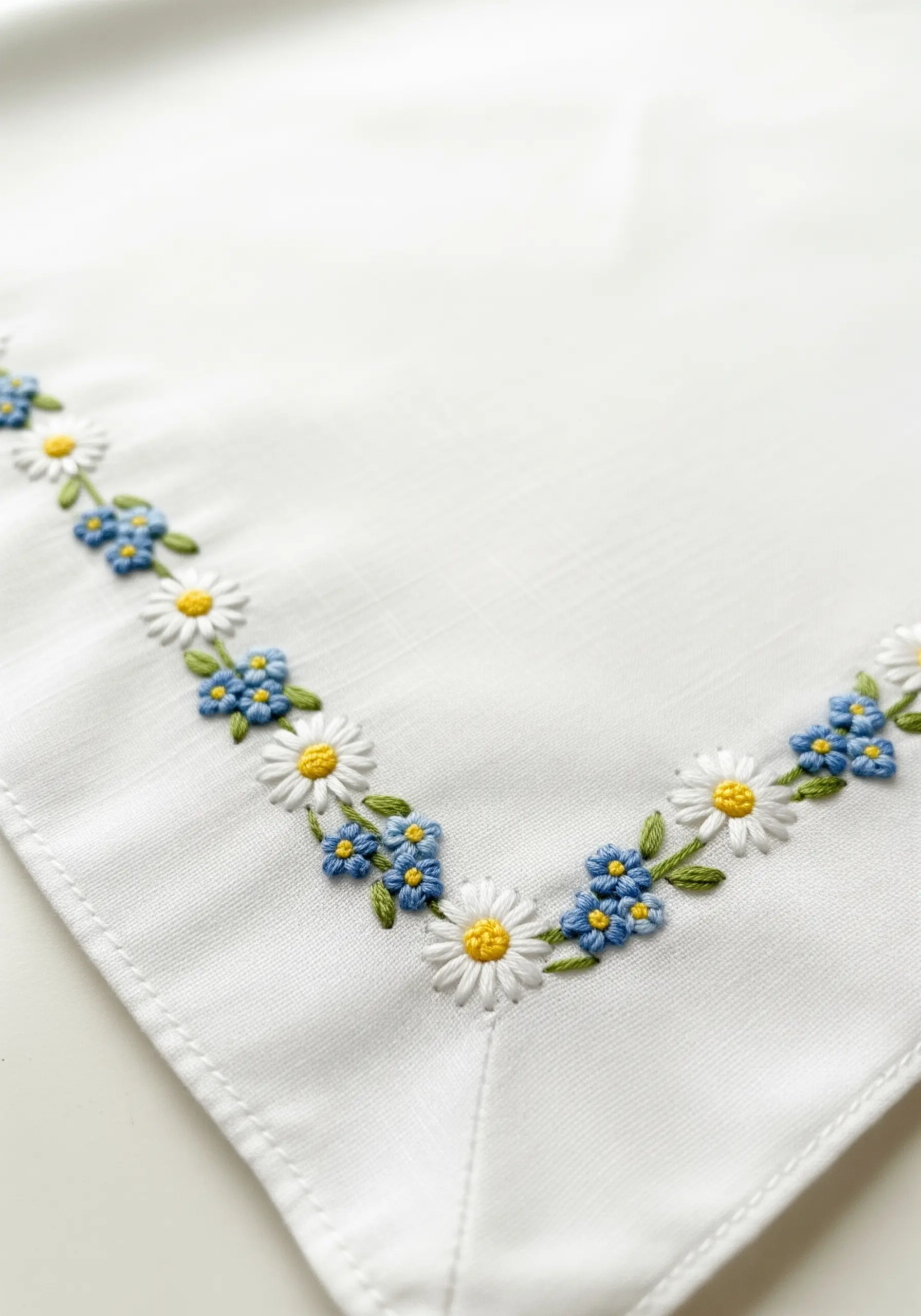 A delicate corner border of embroidered daisies and forget-me-nots on a white napkin.
