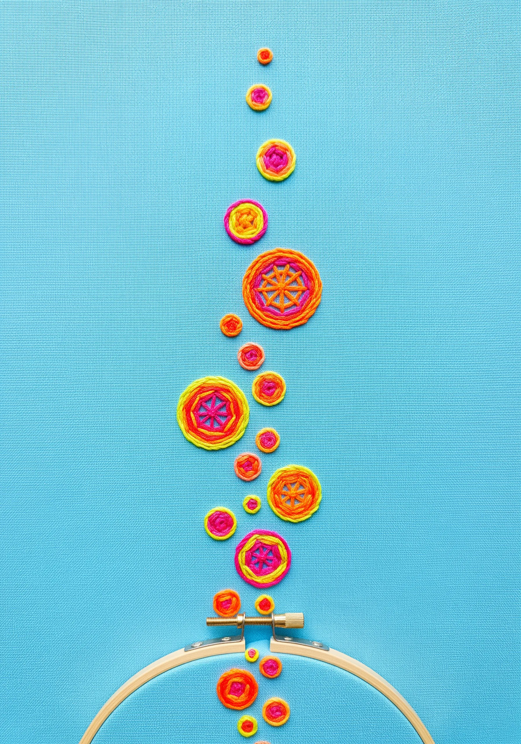 Colorful woven wheel stitches in pink and orange float upwards from the bottom of a hoop.