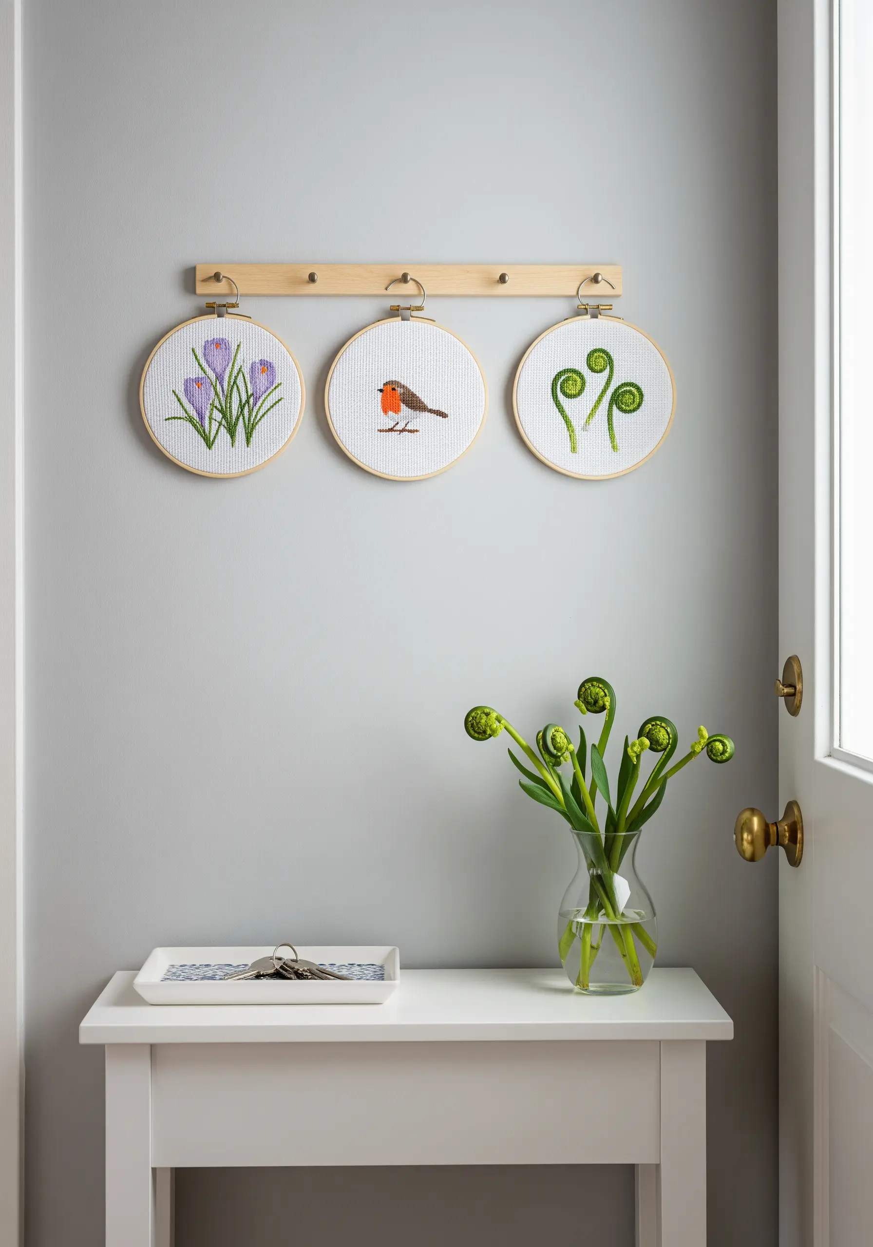 Three cross-stitch embroidery hoops with spring motifs hanging on a wooden rack in an entryway.