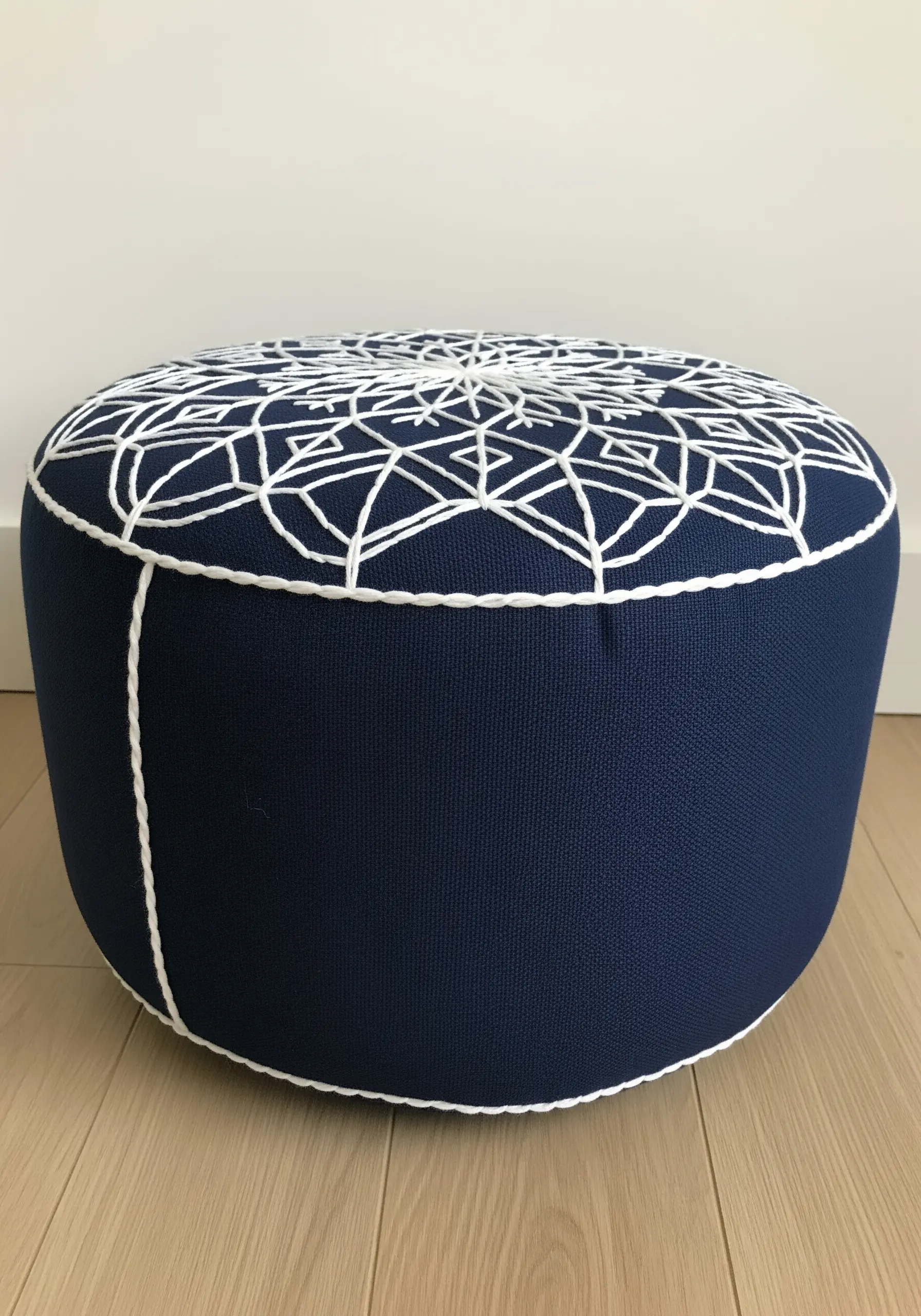 A navy blue ottoman pouf embellished with a large, white geometric mandala pattern in thick cord.
