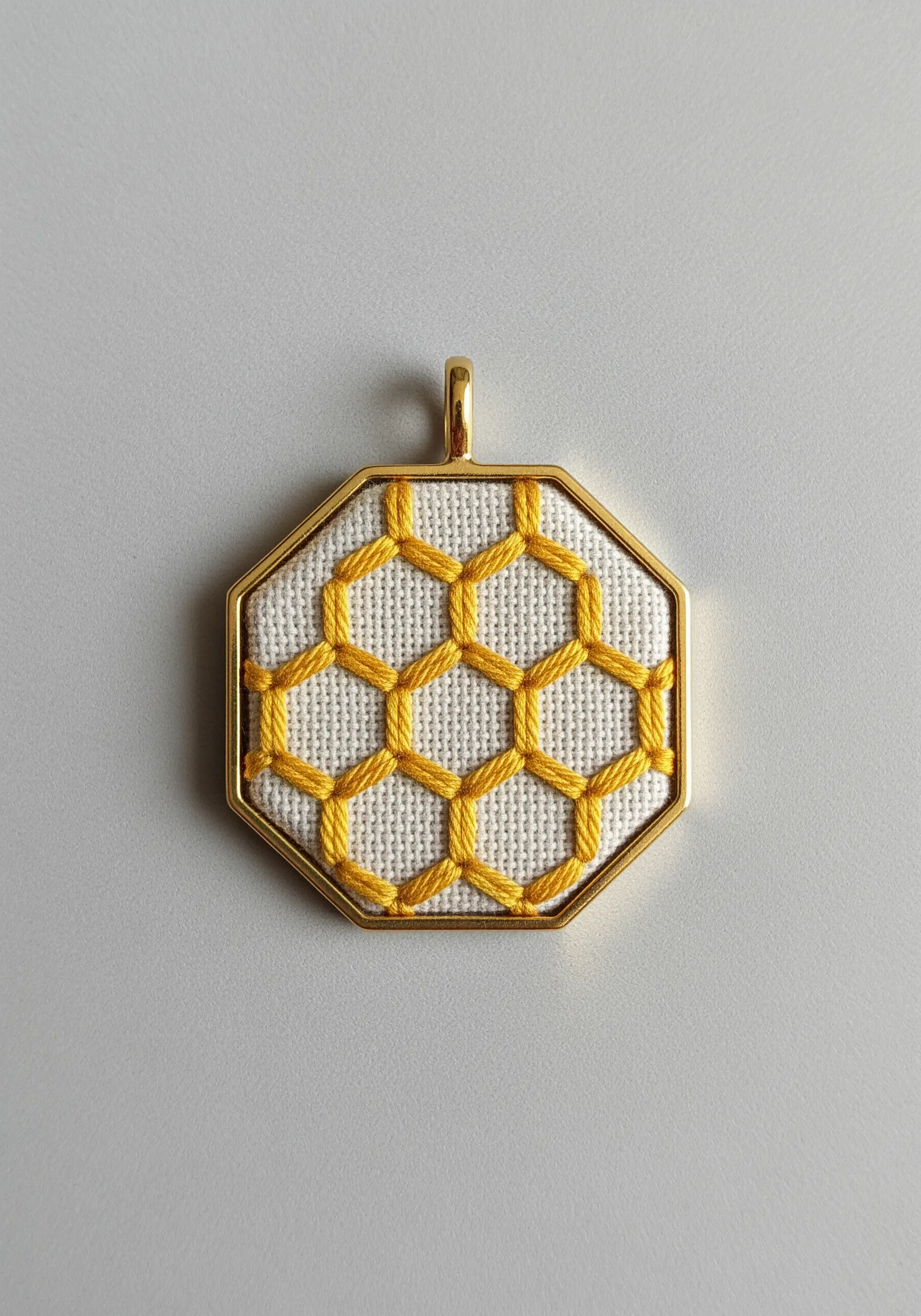 A geometric honeycomb pattern outlined in yellow thread in an octagonal gold pendant.