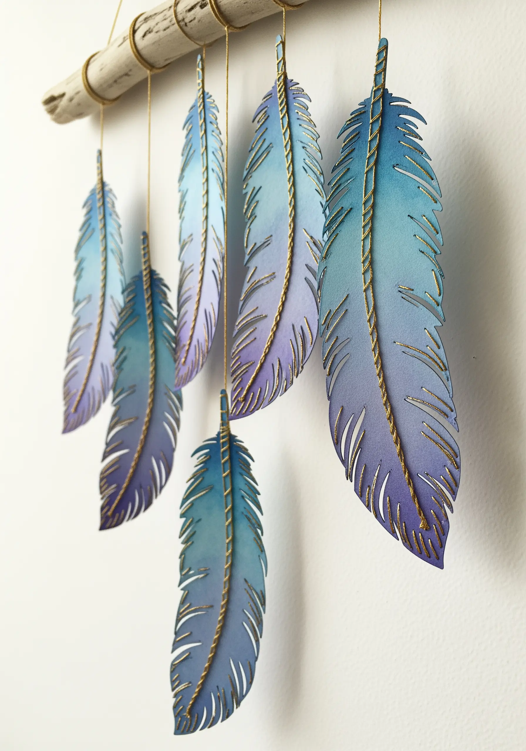 Paper feathers painted in blue and purple gradients, hanging from a branch with gold thread spines.