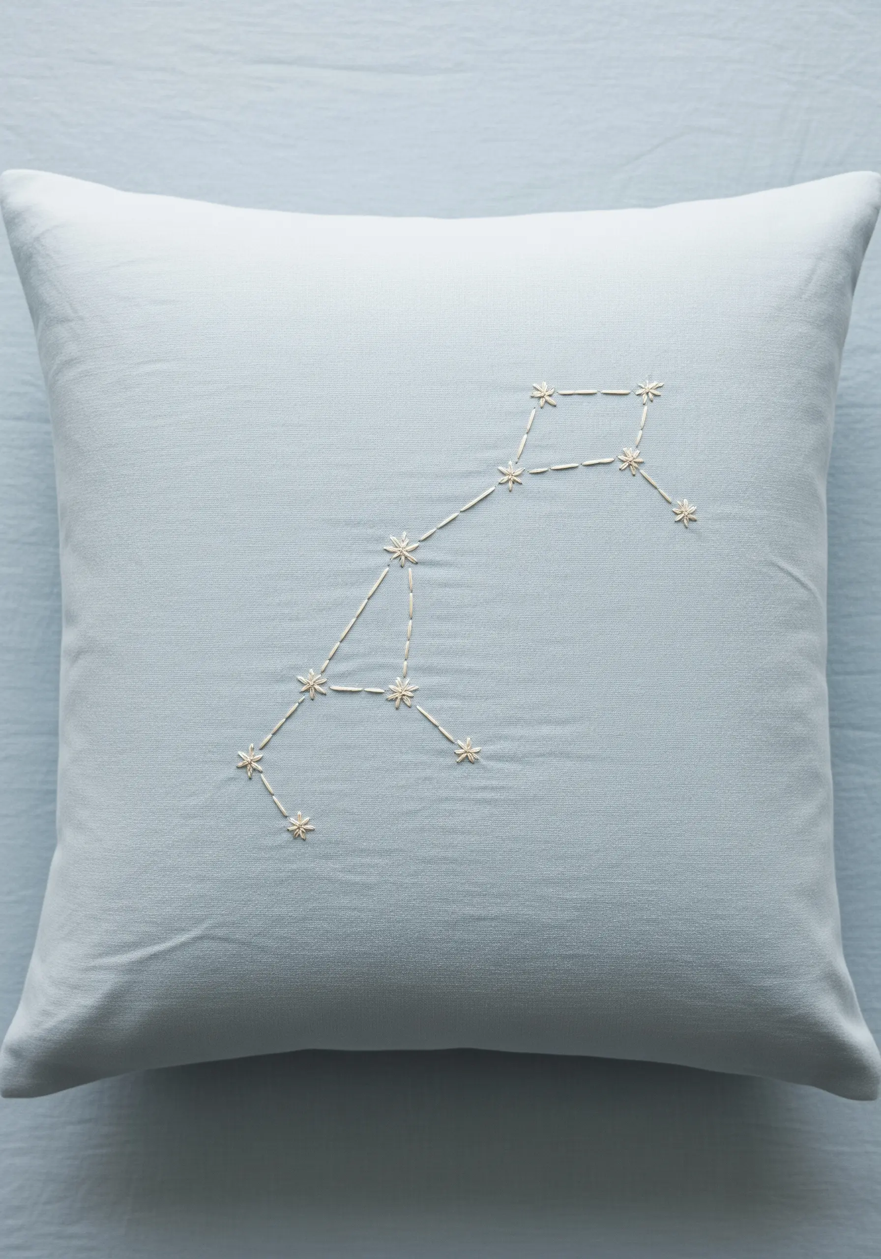 An embroidered constellation using backstitch and star stitches on a light blue pillow.