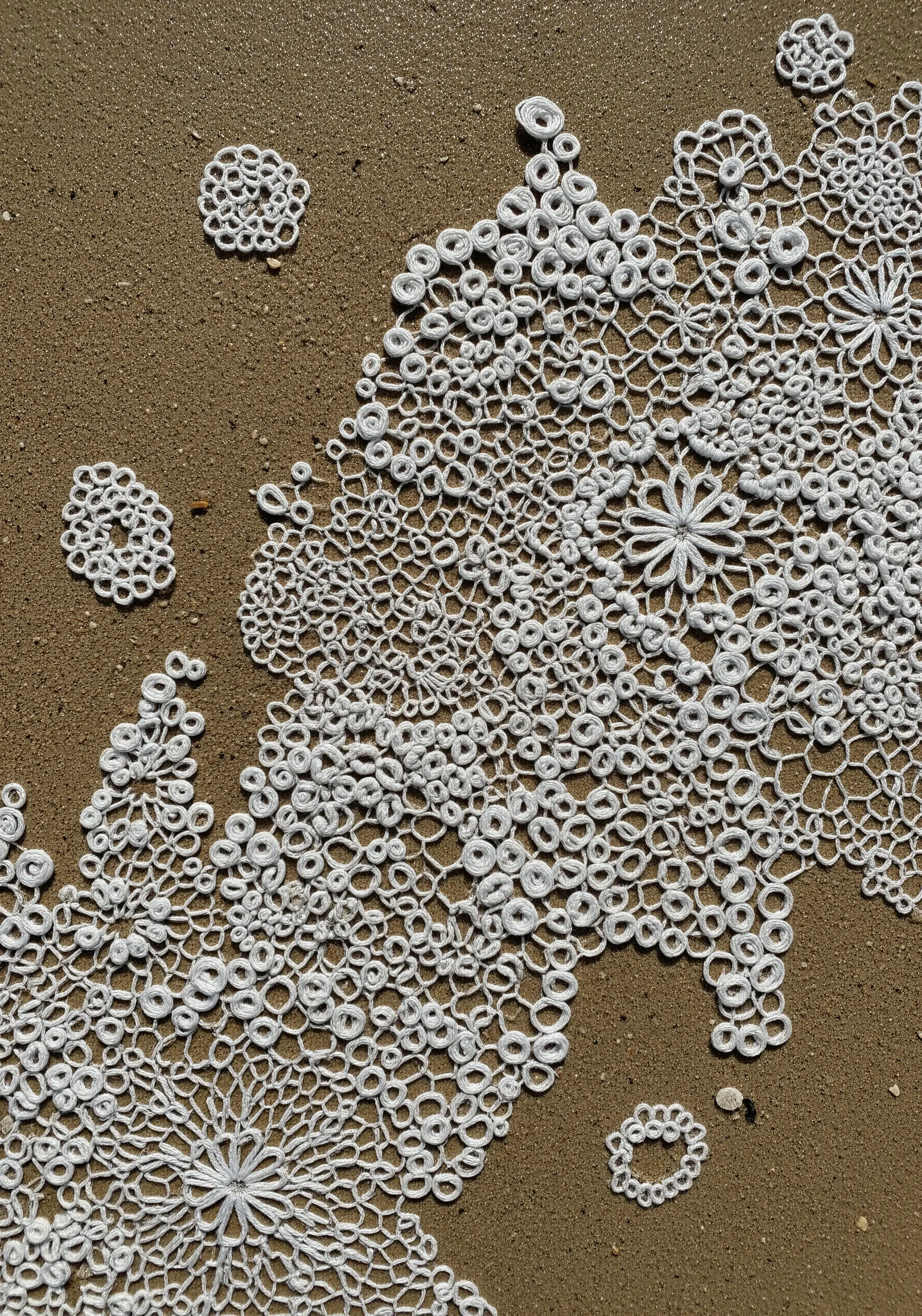 Intricate white lace-like embroidery resembling sea foam, arranged on a sandy surface.