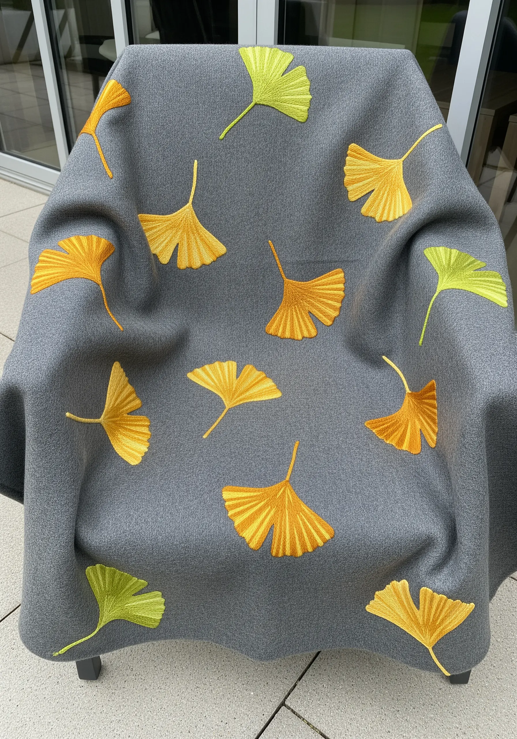 A grey wool throw blanket adorned with scattered embroidered ginkgo leaves in yellow and green.