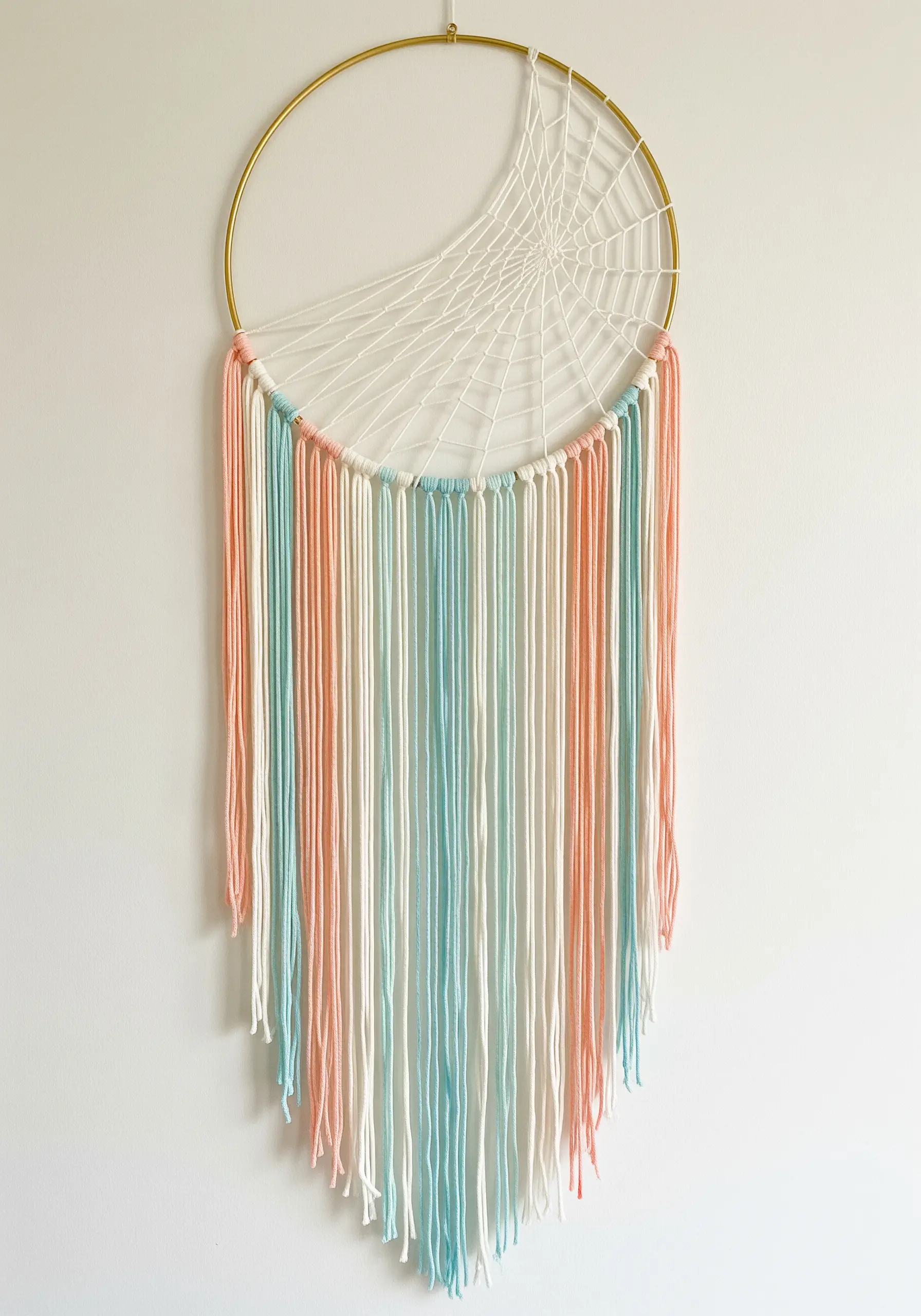 A modern dreamcatcher on a gold hoop with a white crescent web and a long pastel fringe.