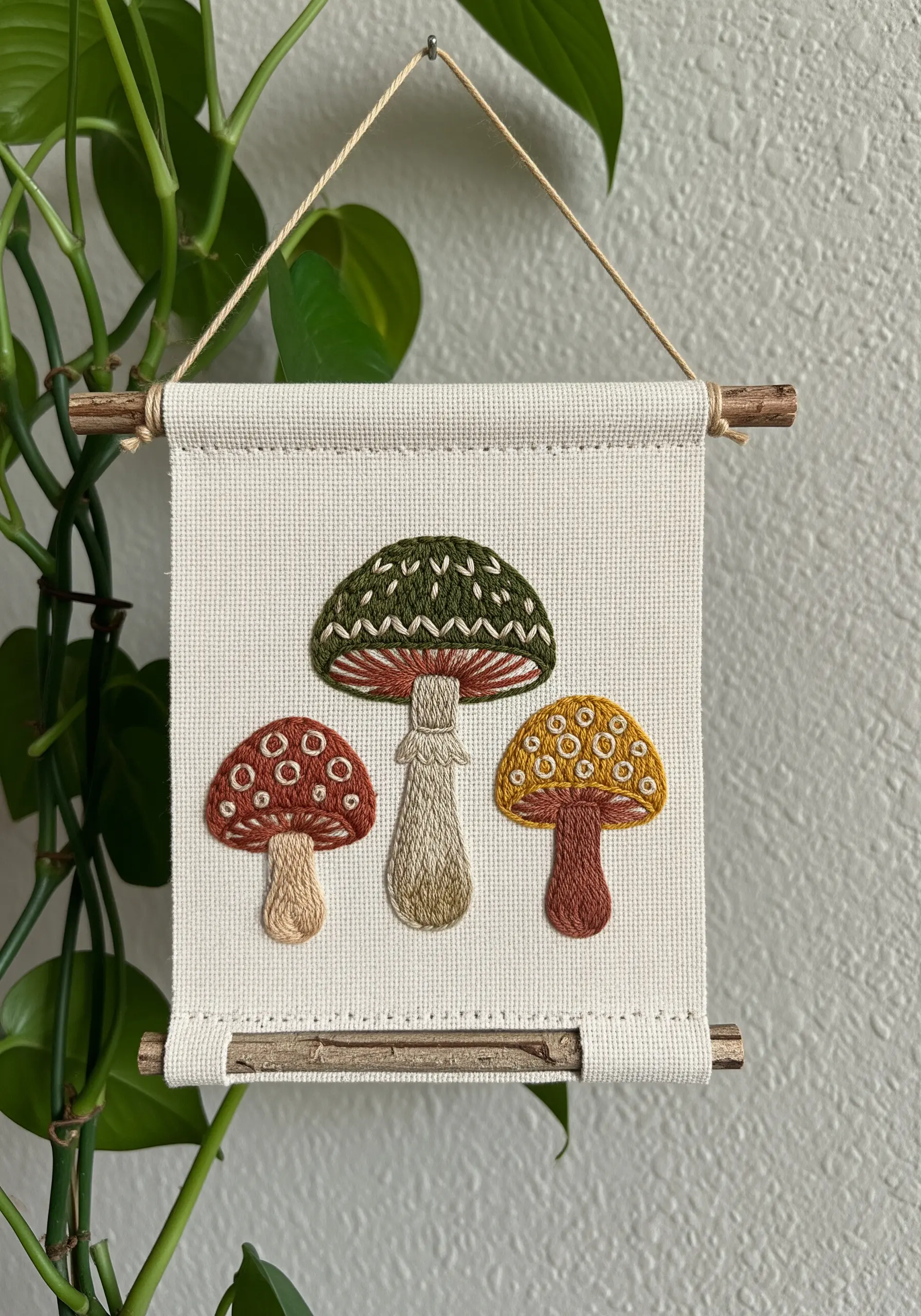 A small wall hanging with three embroidered mushrooms in earthy tones, detailed with varied textures.