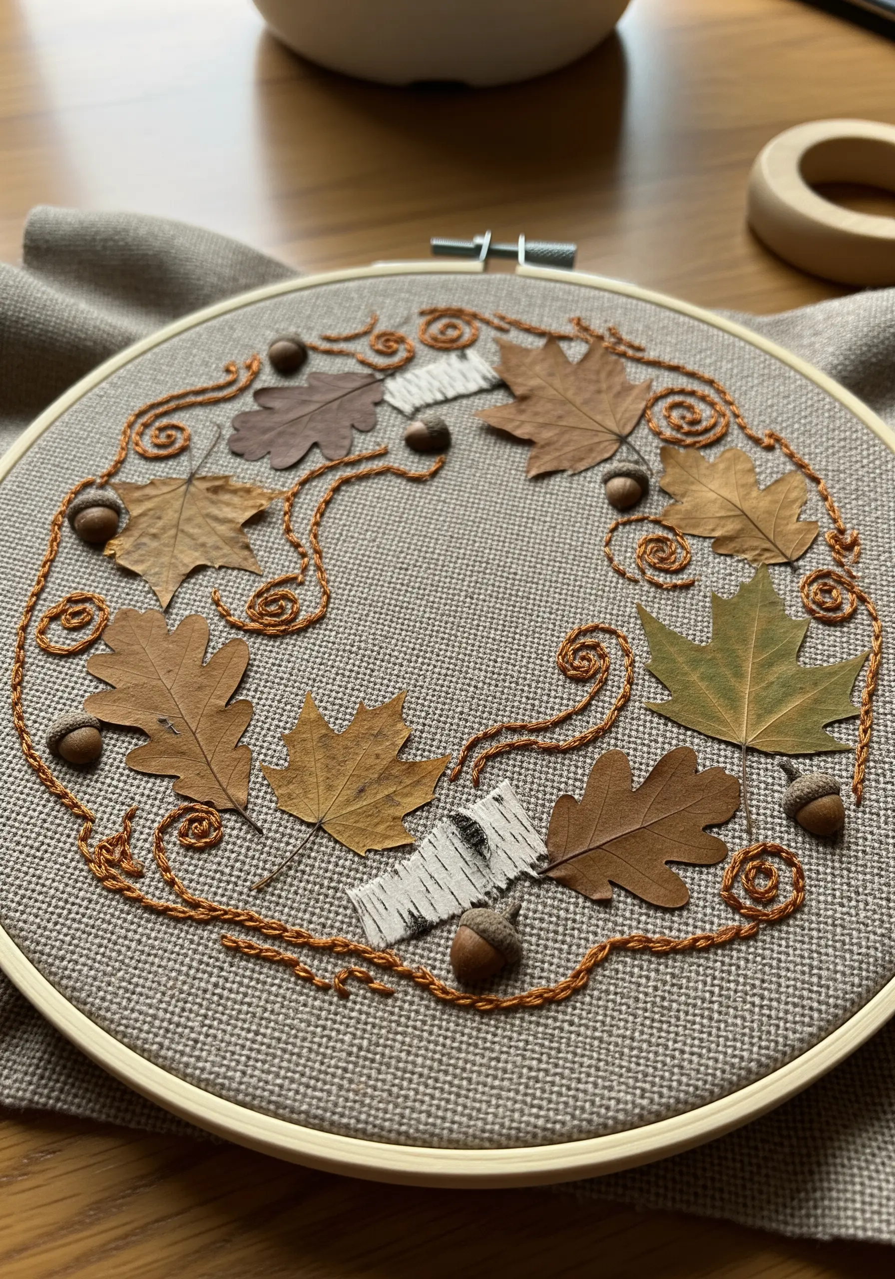An autumnal wreath of pressed leaves, acorns, and birch bark connected by swirling orange embroidery.