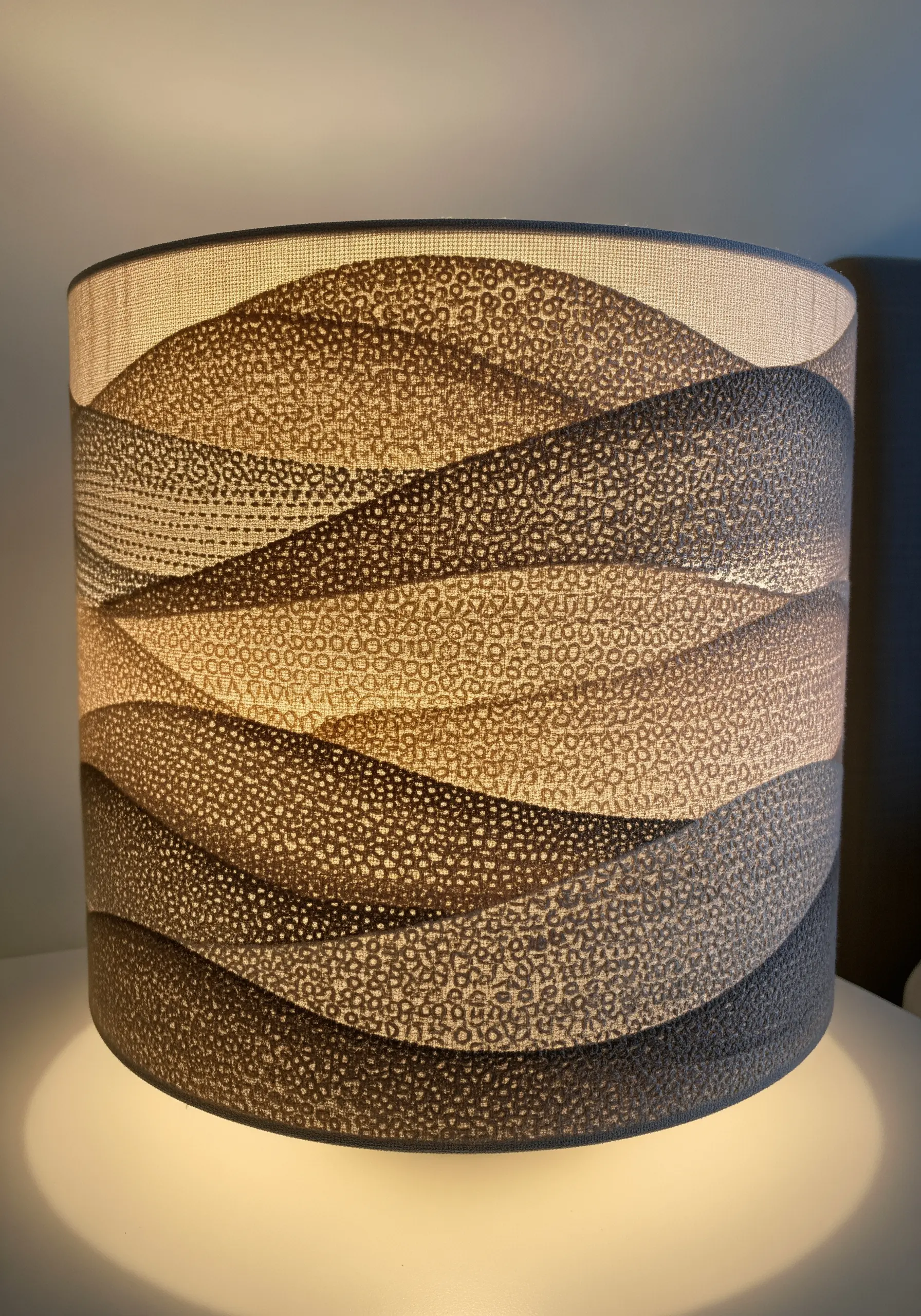 A drum lampshade with an embroidered pattern of wavy hills in various textures and tones.