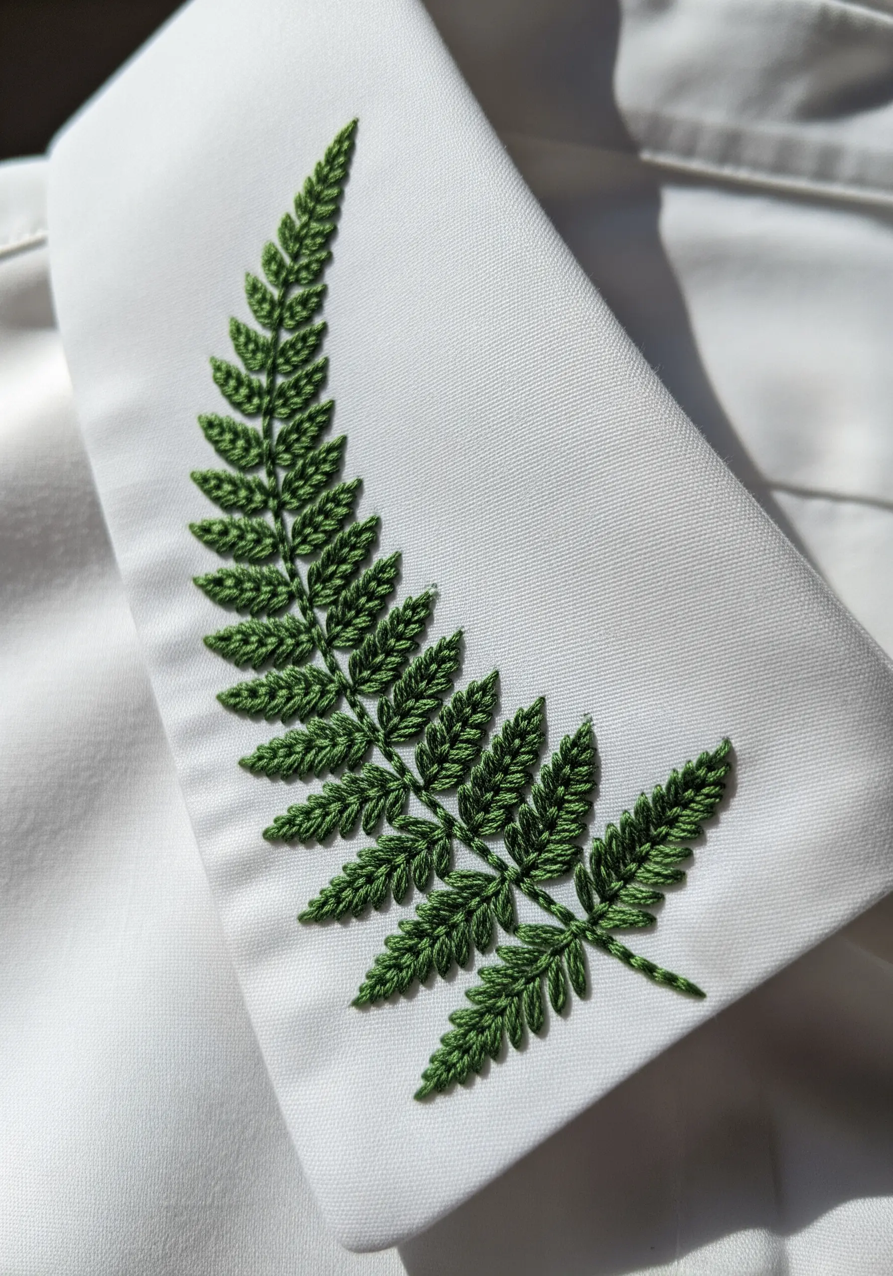 A detailed green fern frond embroidered on a white shirt collar using fishbone stitch.