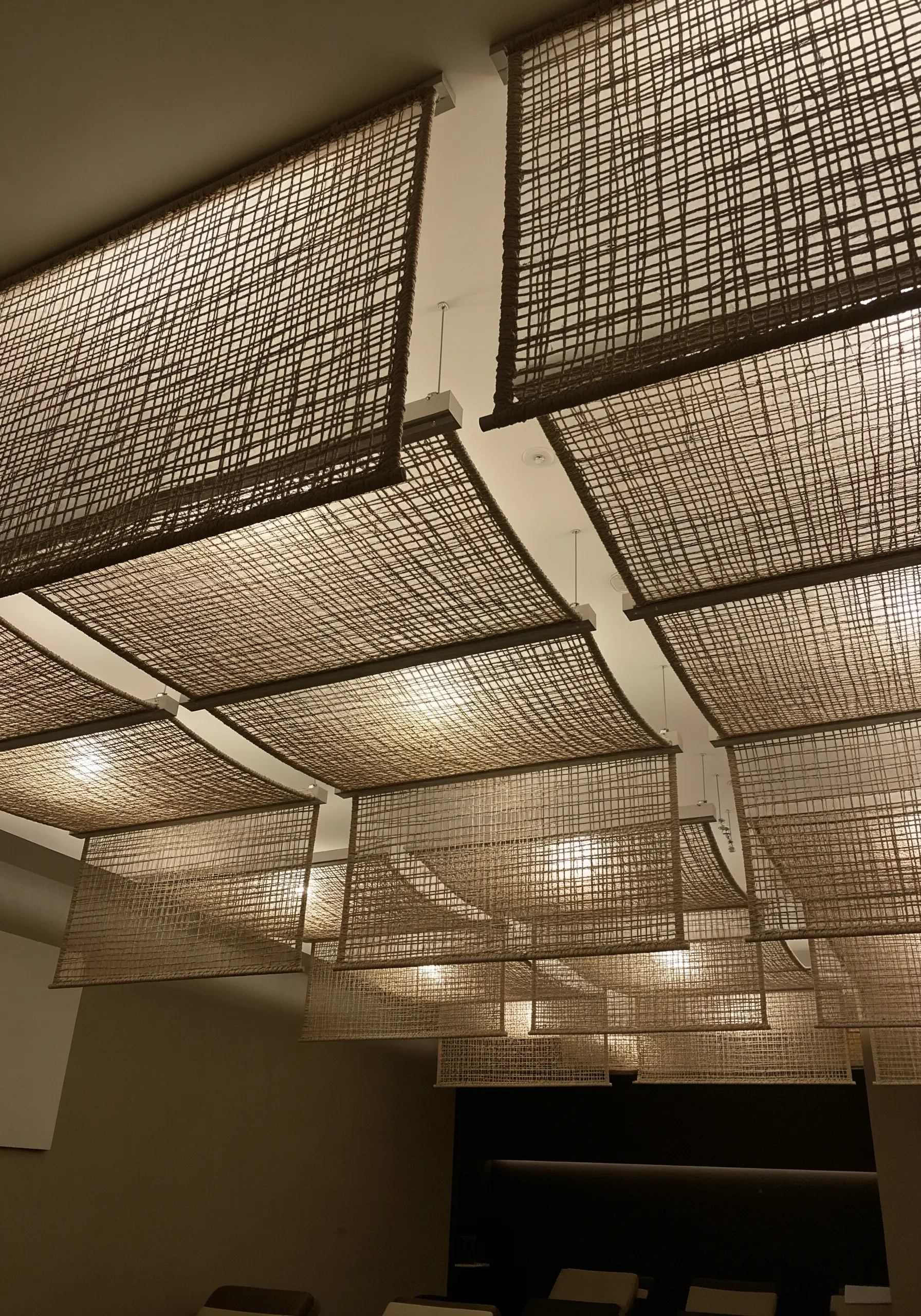 A series of rectangular, open-weave panels made of dark fiber, suspended from a ceiling to diffuse light.