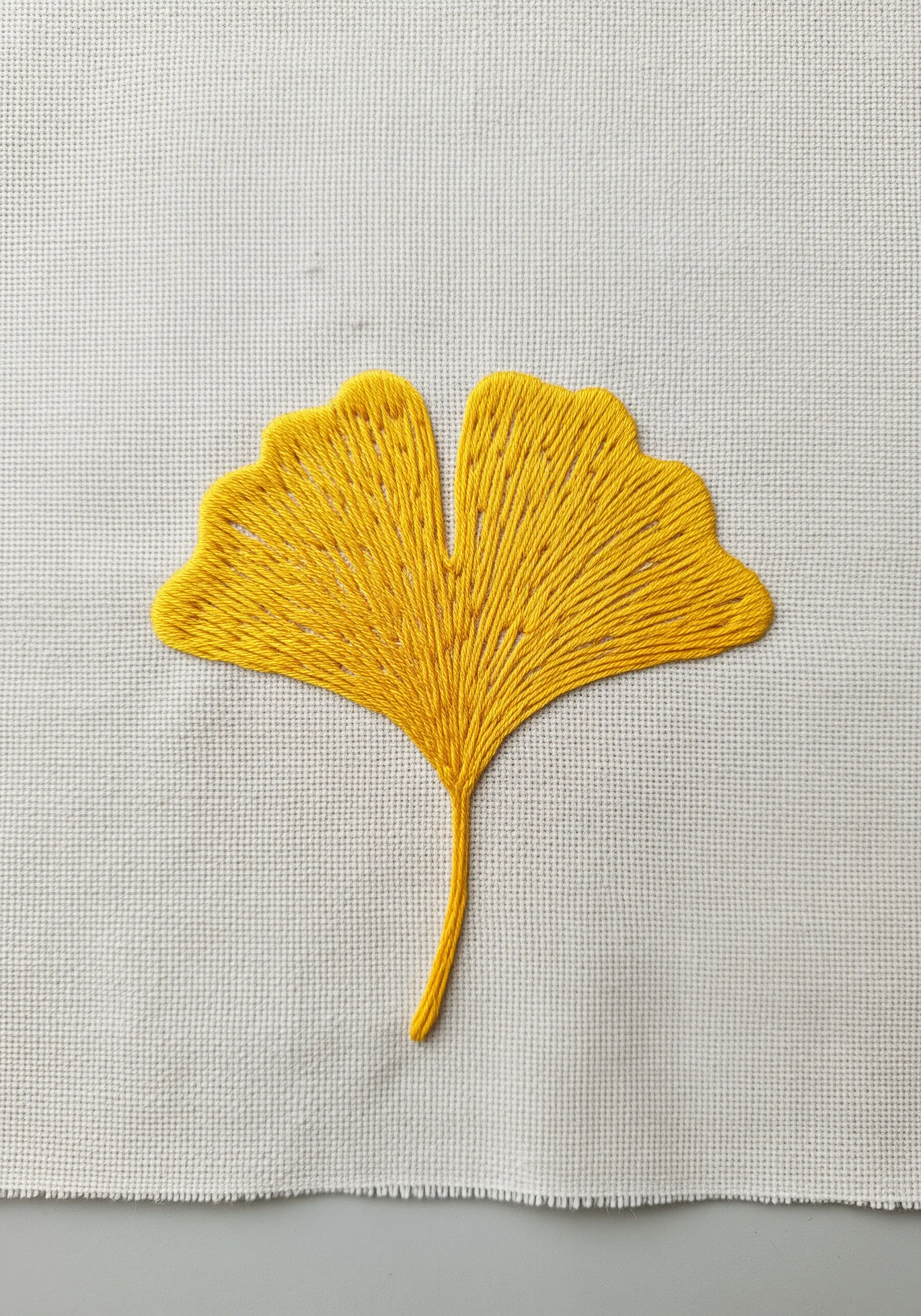 A bright yellow ginkgo leaf embroidered with directional satin stitch on white Aida.