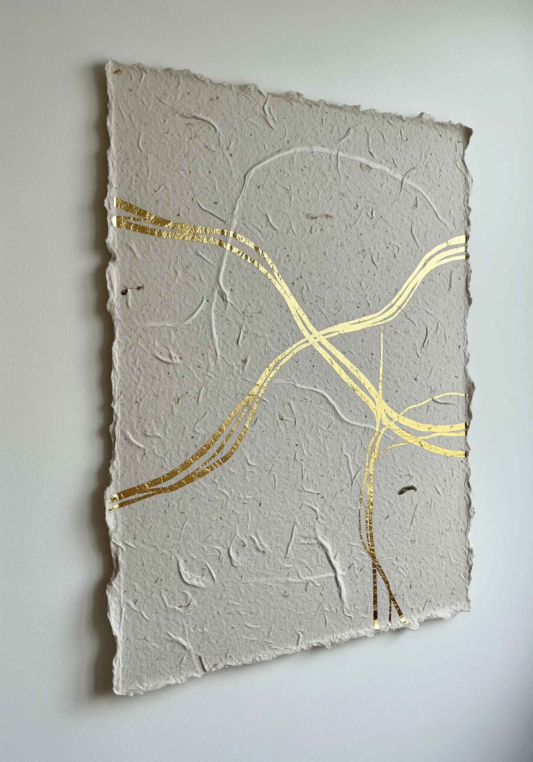 Abstract intersecting gold leaf lines on a sheet of rough, deckle-edge handmade paper.