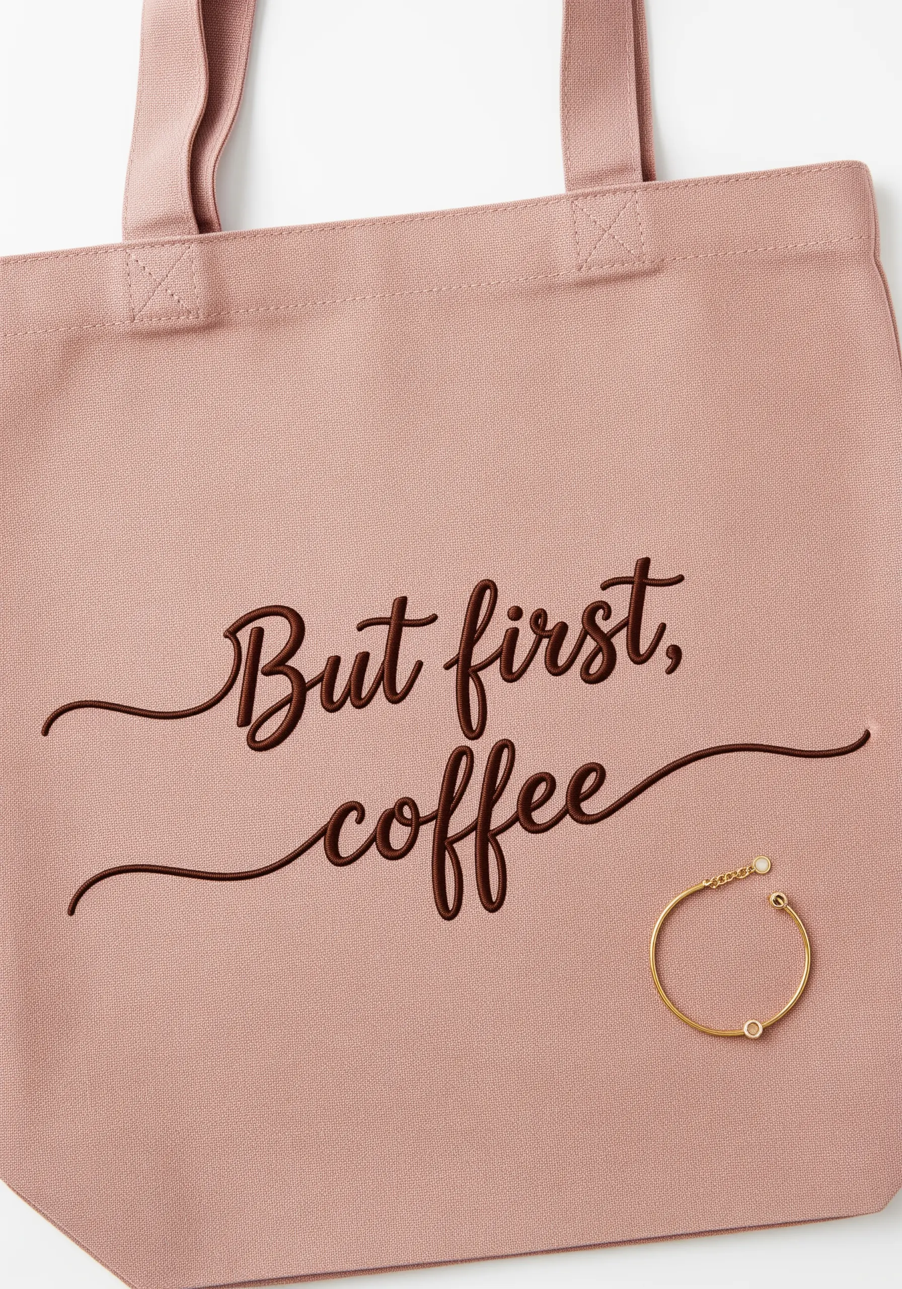 Embroidered script text 'But first, coffee' in a flowing brown font on a pink tote bag.