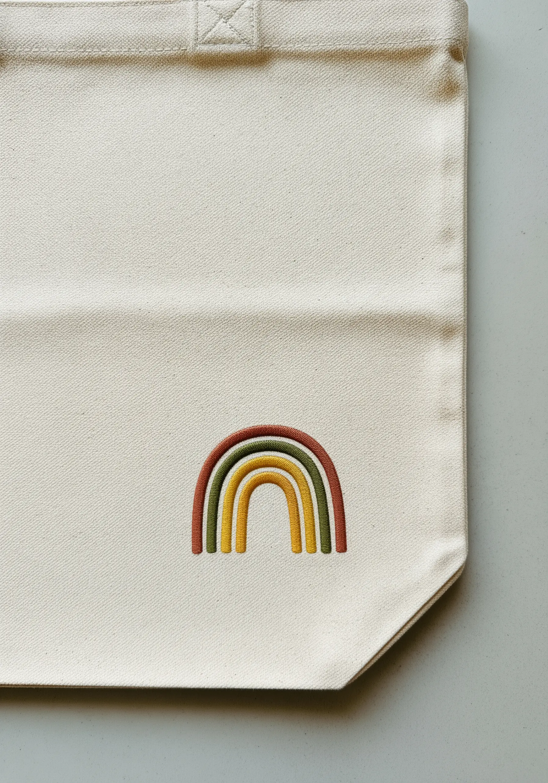 A small embroidered rainbow in muted earth tones on the corner of a canvas tote.