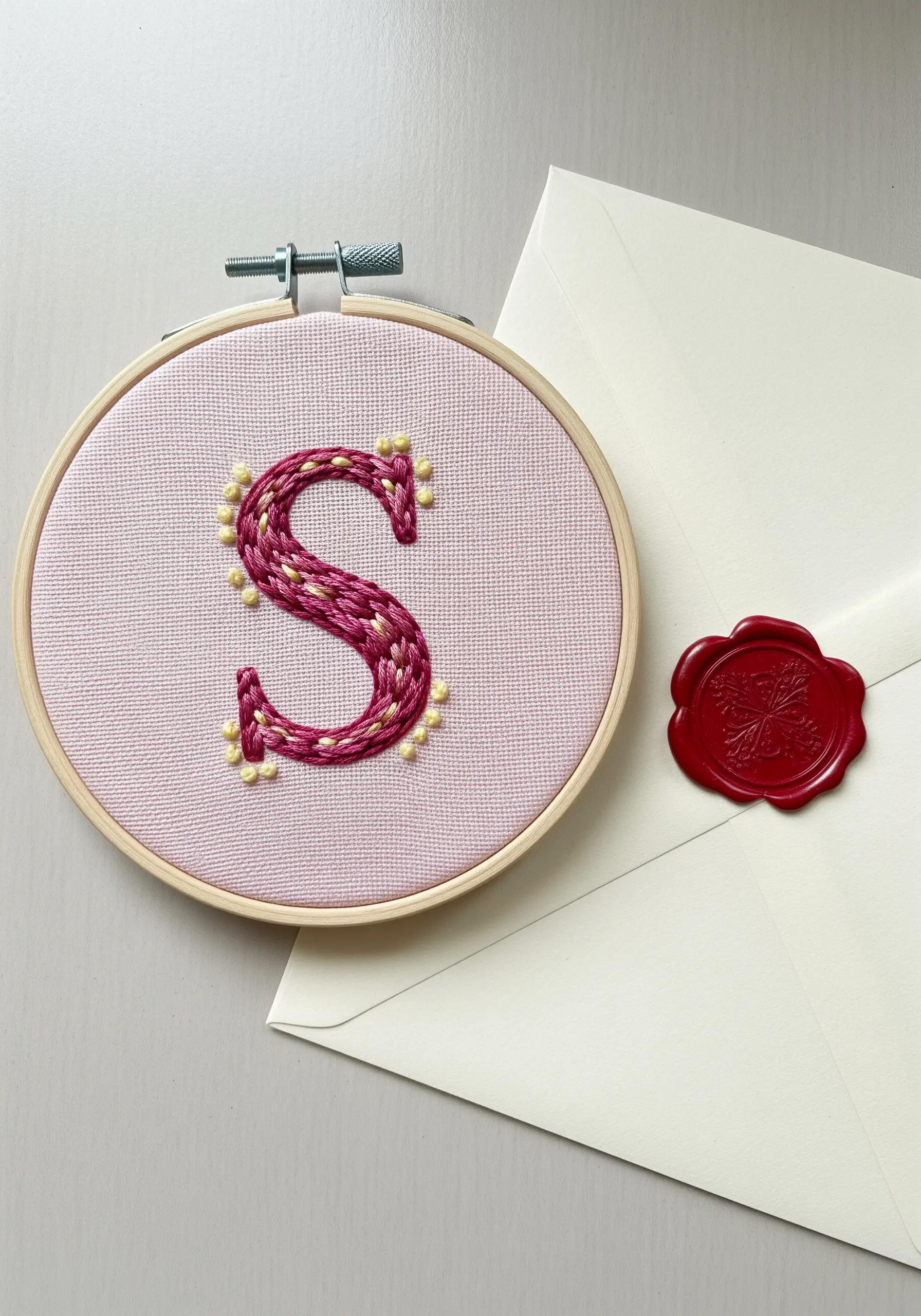 An embroidered letter 'S' with a woven texture and pearl-like French knot details.