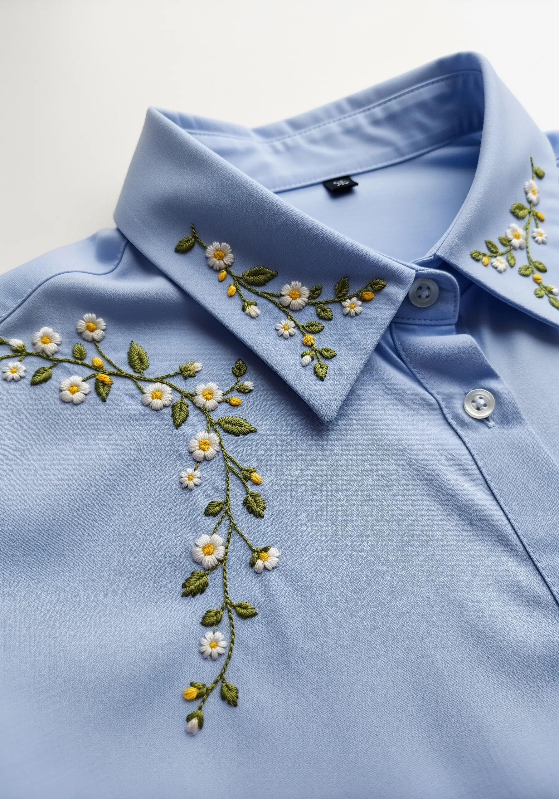 Delicate daisy vines embroidered asymmetrically on the collar of a light blue button-down shirt.