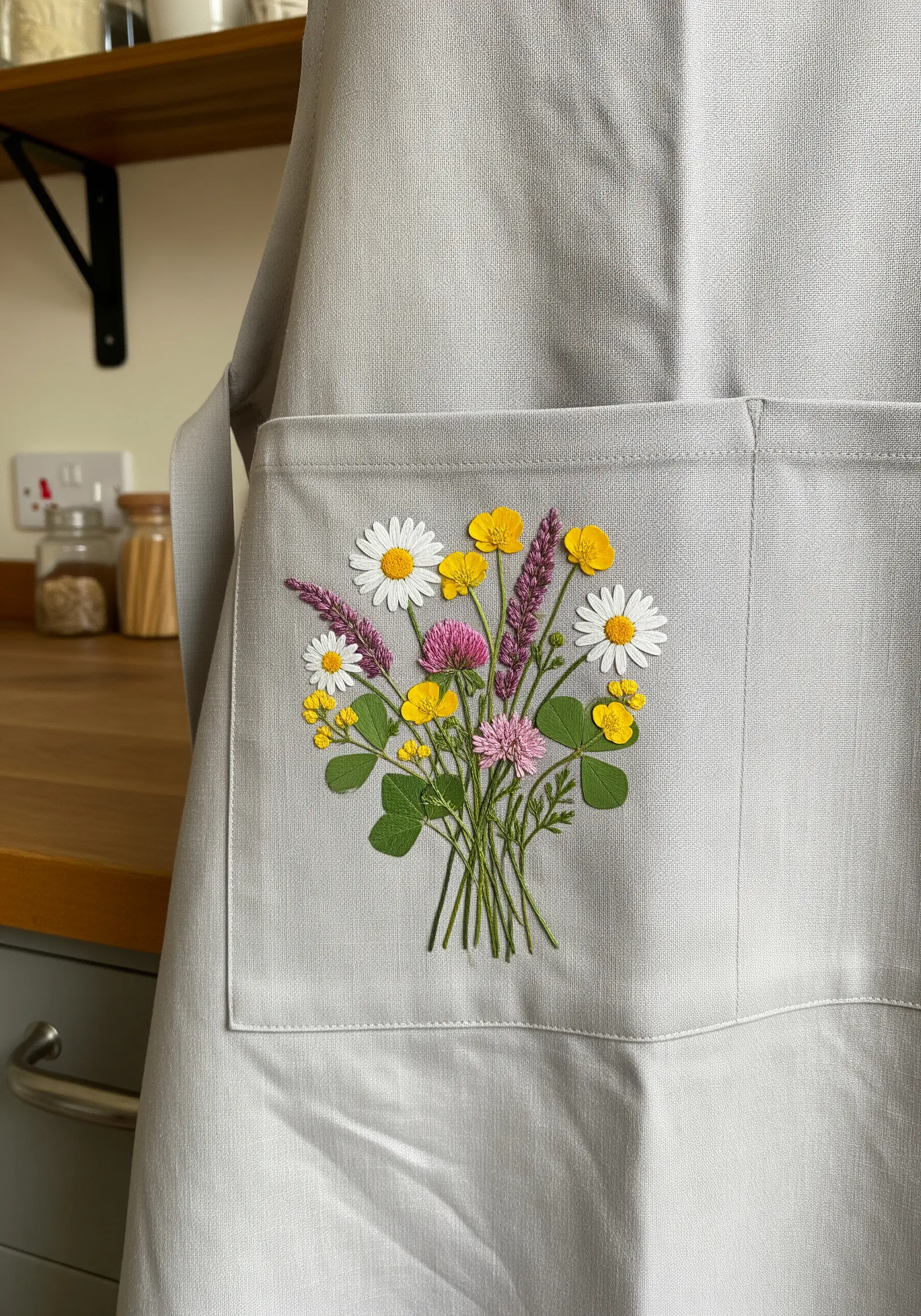 A detailed, colorful bouquet of embroidered wildflowers stitched onto the pocket of a gray apron.