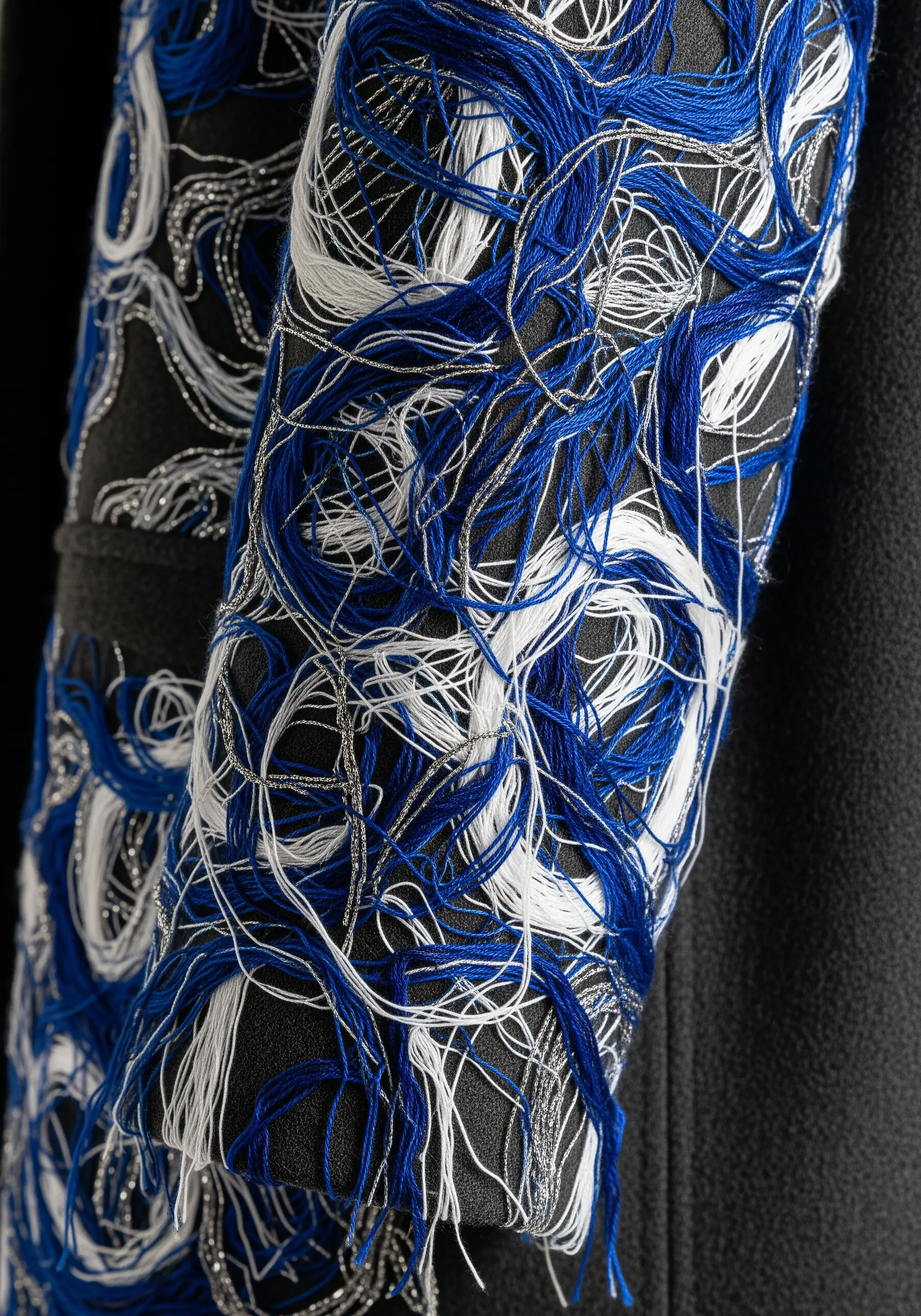 A close-up of a black sleeve embellished with chaotic swirls of royal blue, white, and silver thread