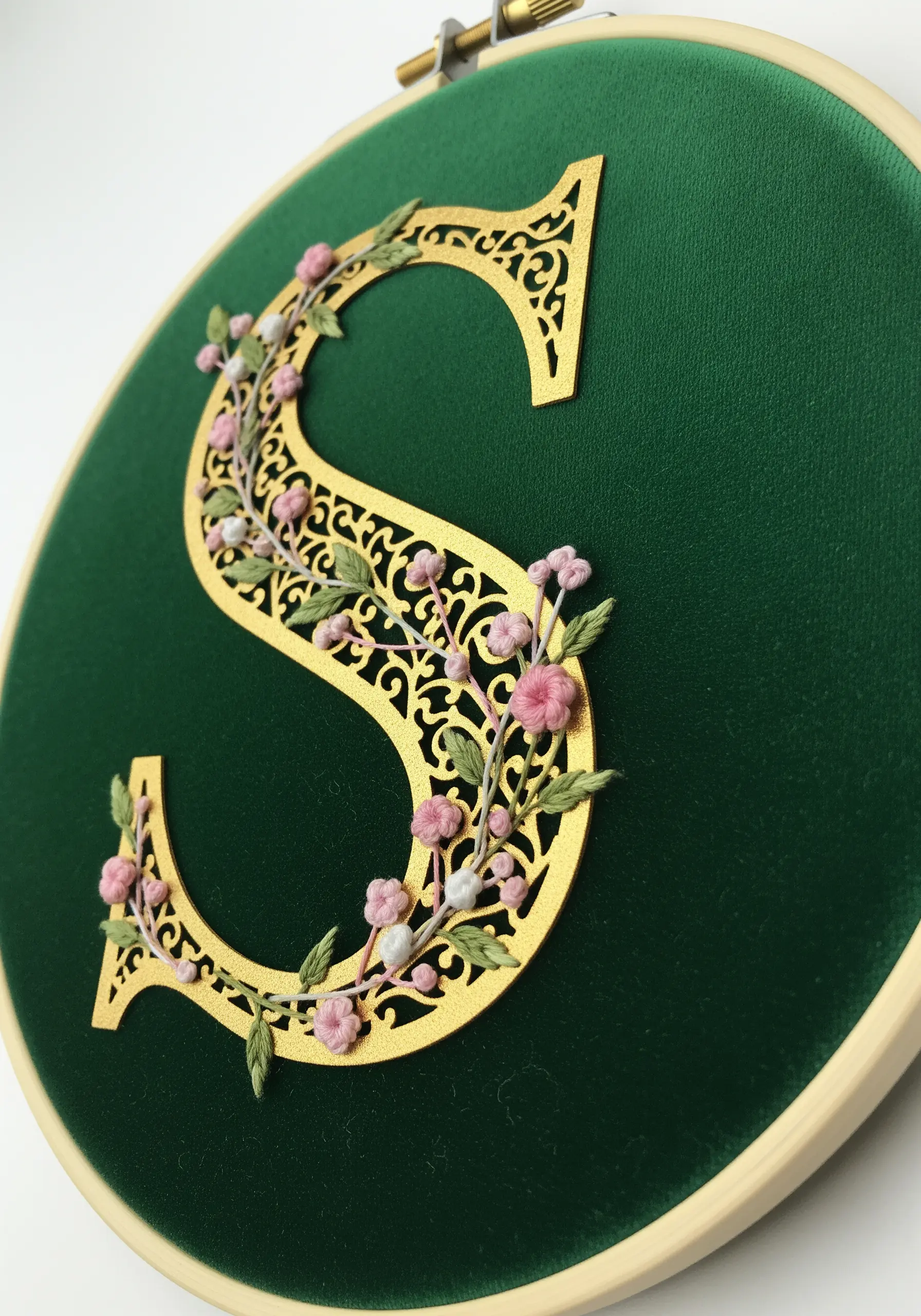 A gold filigree paper letter 'S' on green fabric, embellished with tiny pink and white embroidered flowers.