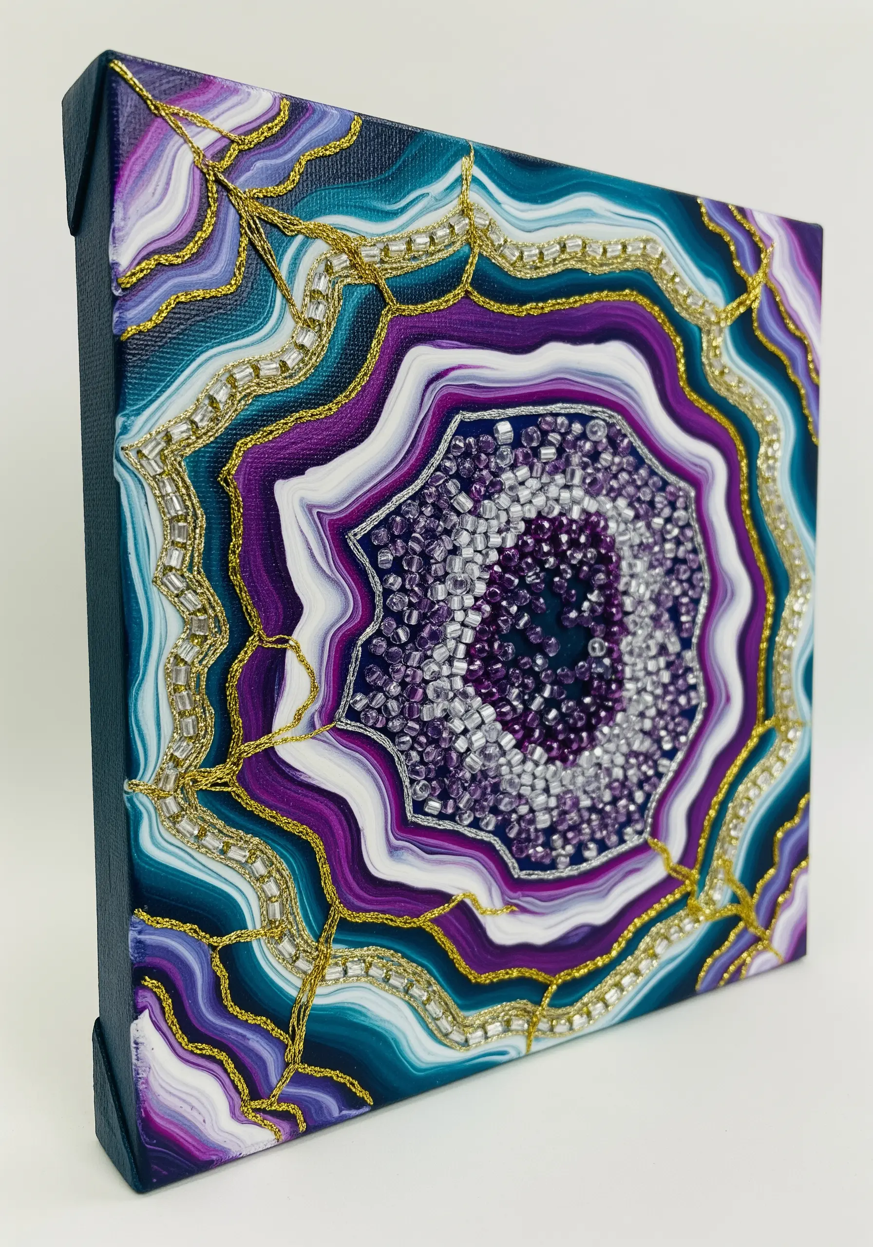 A painted geode on canvas, with the layers outlined in gold thread and the center filled with beads.