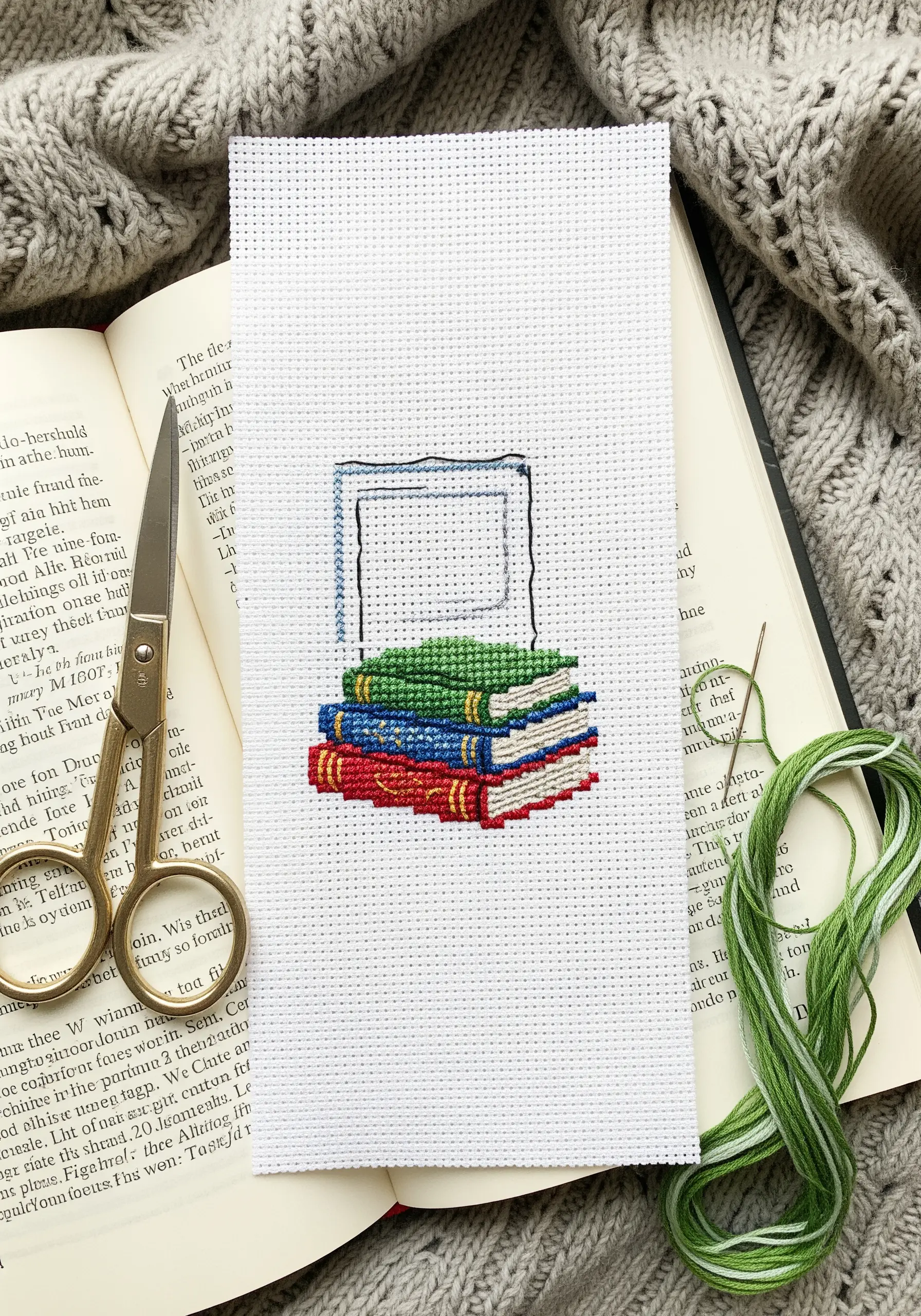 Cross-stitch design of a stack of colorful books on white Aida cloth.